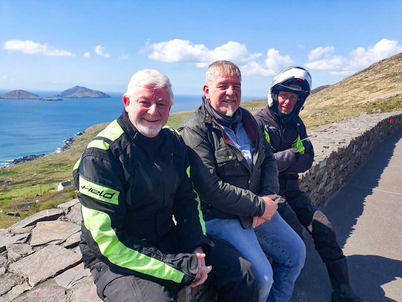 Three smiling motorcyclists resting on a stone wall above the sea at Coomakista Pass, Ring of Kerry