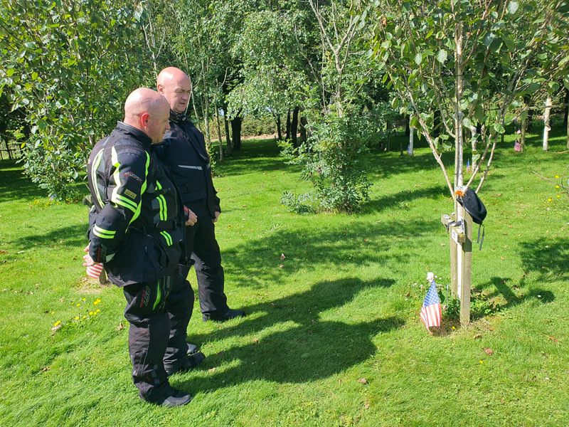 Two motorcyclists pausing at a memorial tree with an American flag in the Ringfinnan Garden of Remembrance near Kinsale