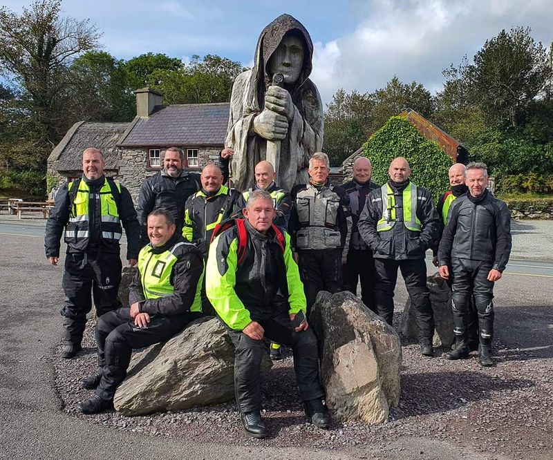 A Wild Atlantic Biker tour group poses at a carved wooden statue beside a stone cottage
