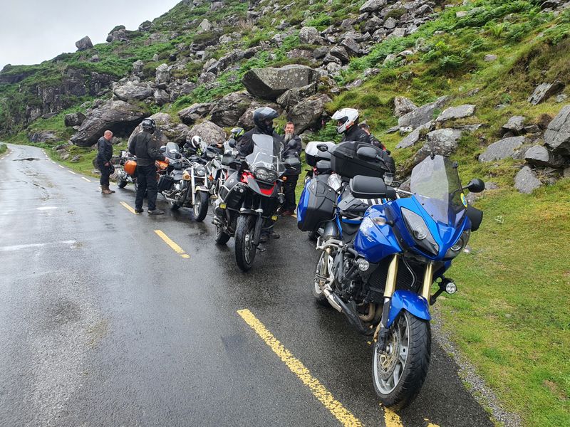 Motorcyclists paused on a wet single-track mountain pass road beneath rocky slopes