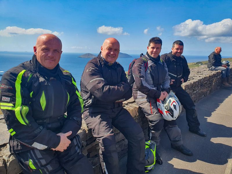 Group of motorcyclists in touring gear resting on a stone wall at a Ring of Kerry coastal viewpoint, islands and blue Atlantic behind them