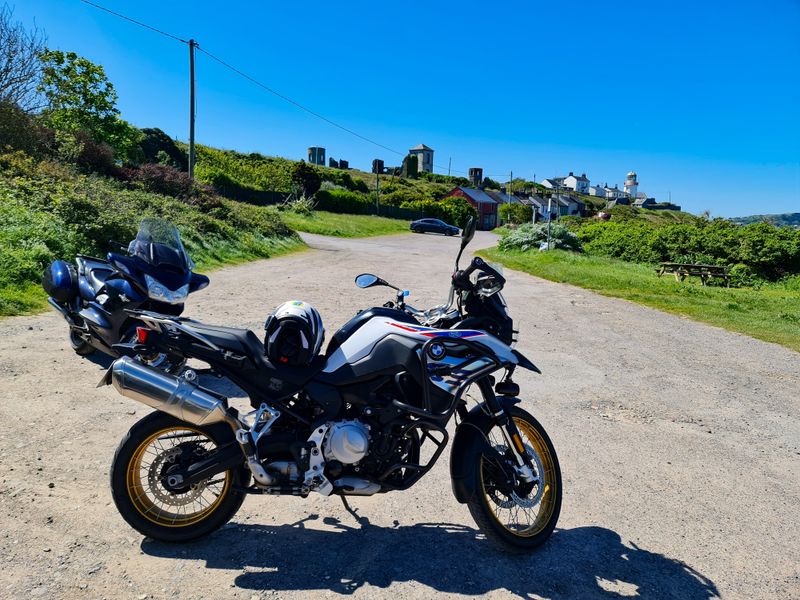 Motorcycles parked on a gravel lane below a coastal village with a white lighthouse under a clear blue sky