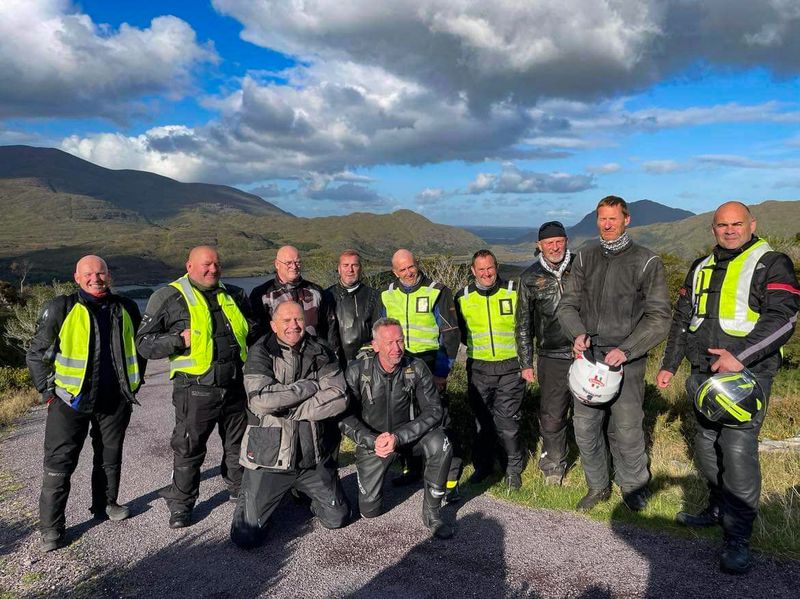 A group of twelve motorcyclists posing together at a scenic Kerry mountain viewpoint