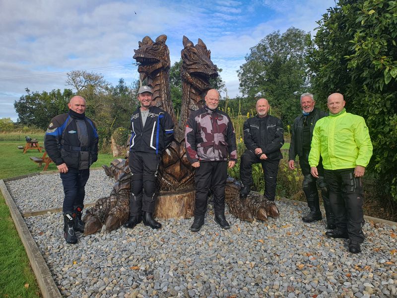 Group of six motorcyclists posing beside a carved wooden dragon sculpture on a tour stop