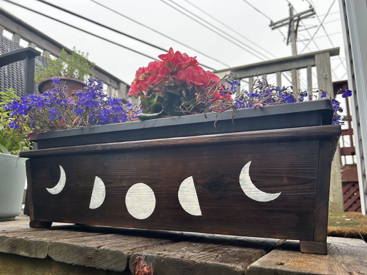 Dark-stained wooden planter box painted with white moon phases, filled with flowers