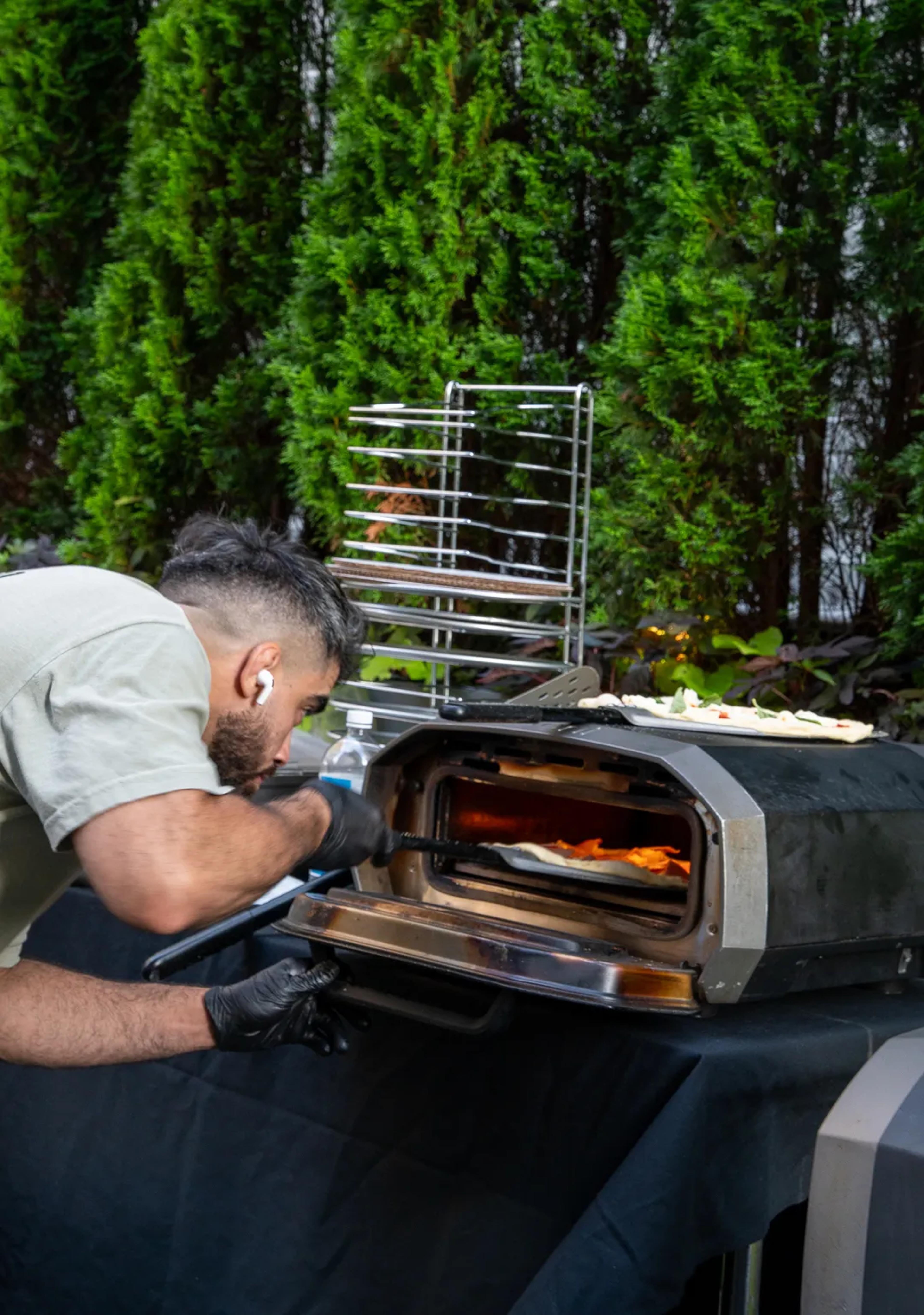A cook bending to check a pie in a countertop oven on the lawn, a pie rack and a clipped hedge behind him