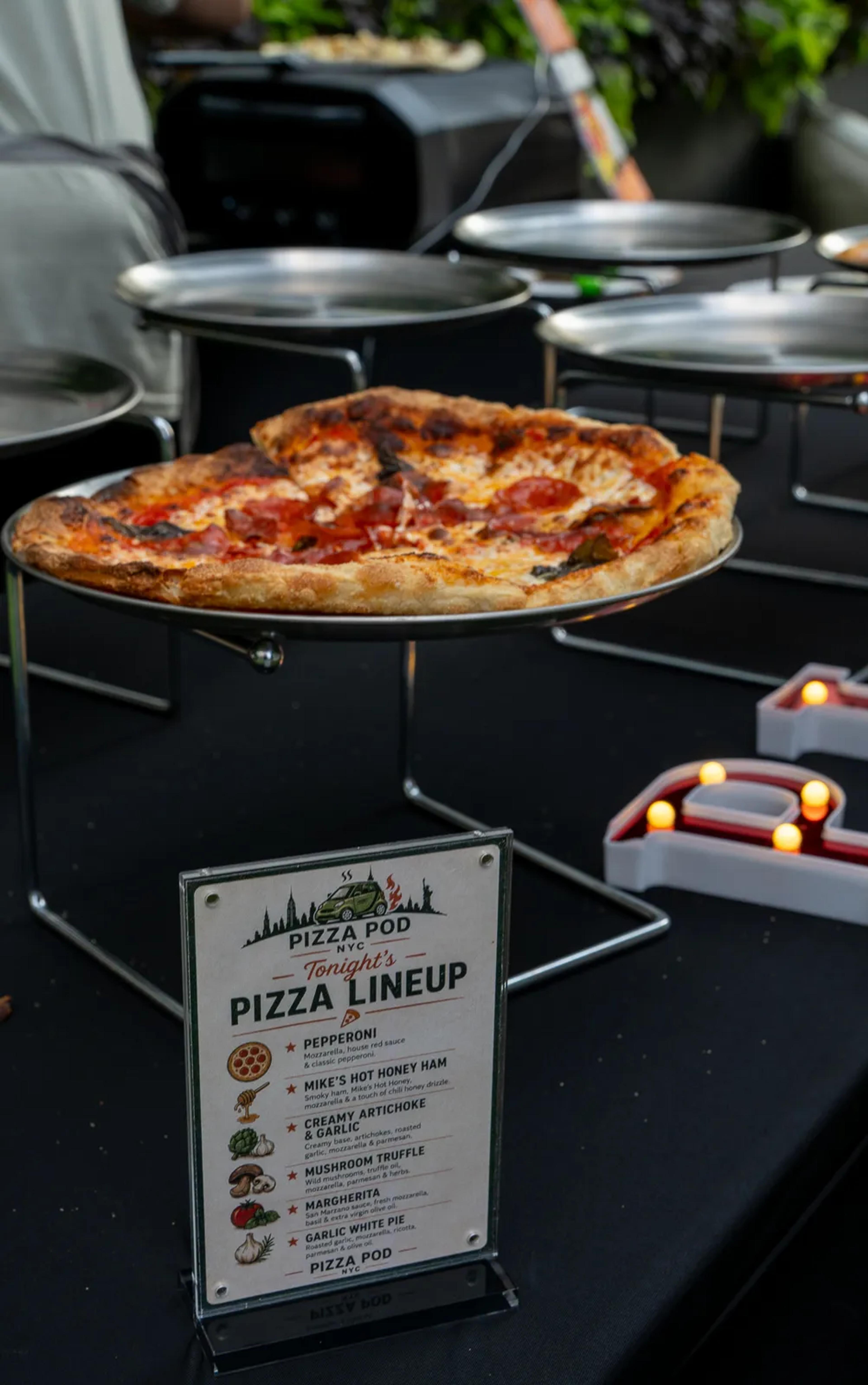 The night's pizza lineup card on the service table, a pepperoni pie on a raised stand behind it