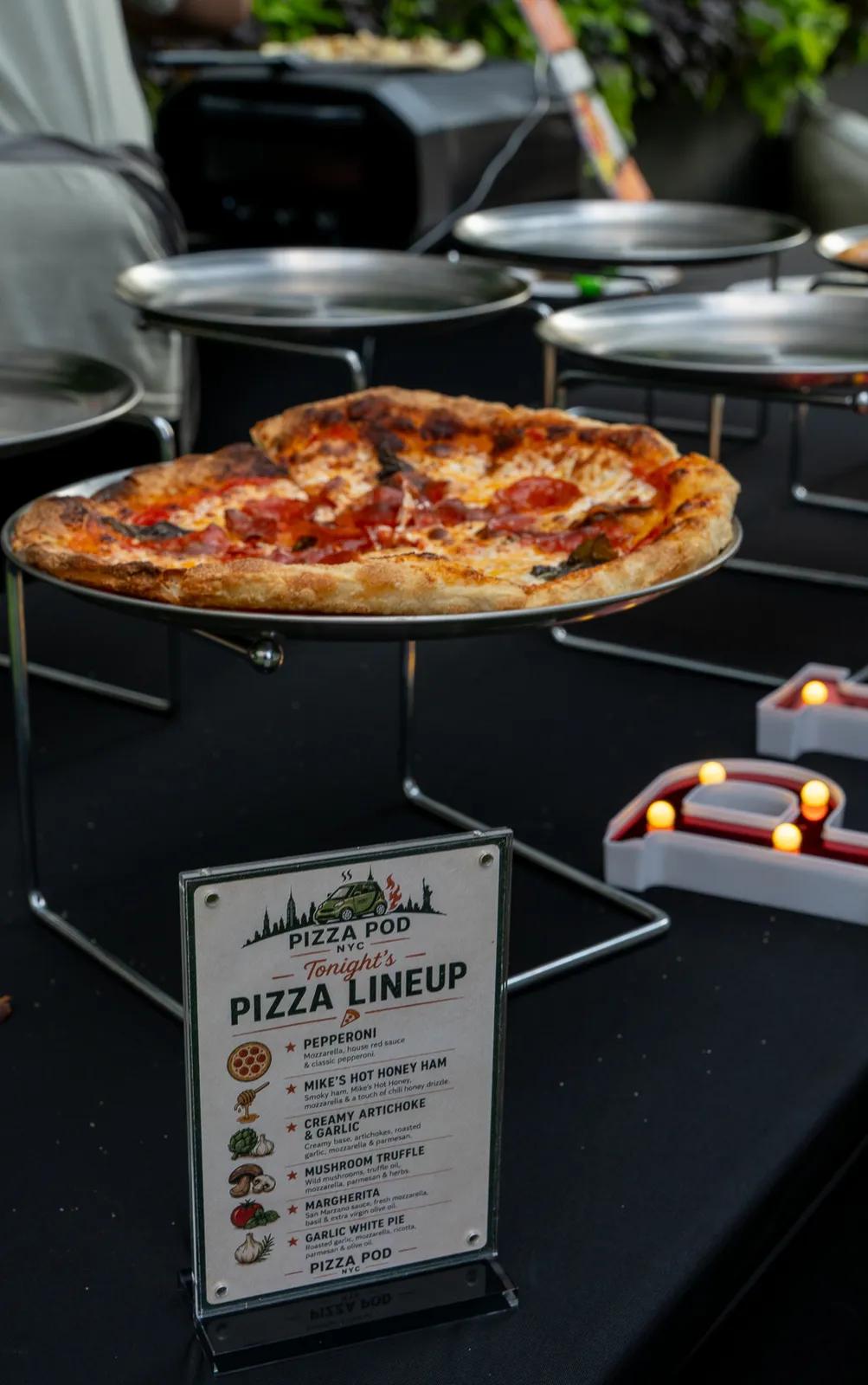 The night's pizza lineup card on the service table, a pepperoni pie on a raised stand behind it