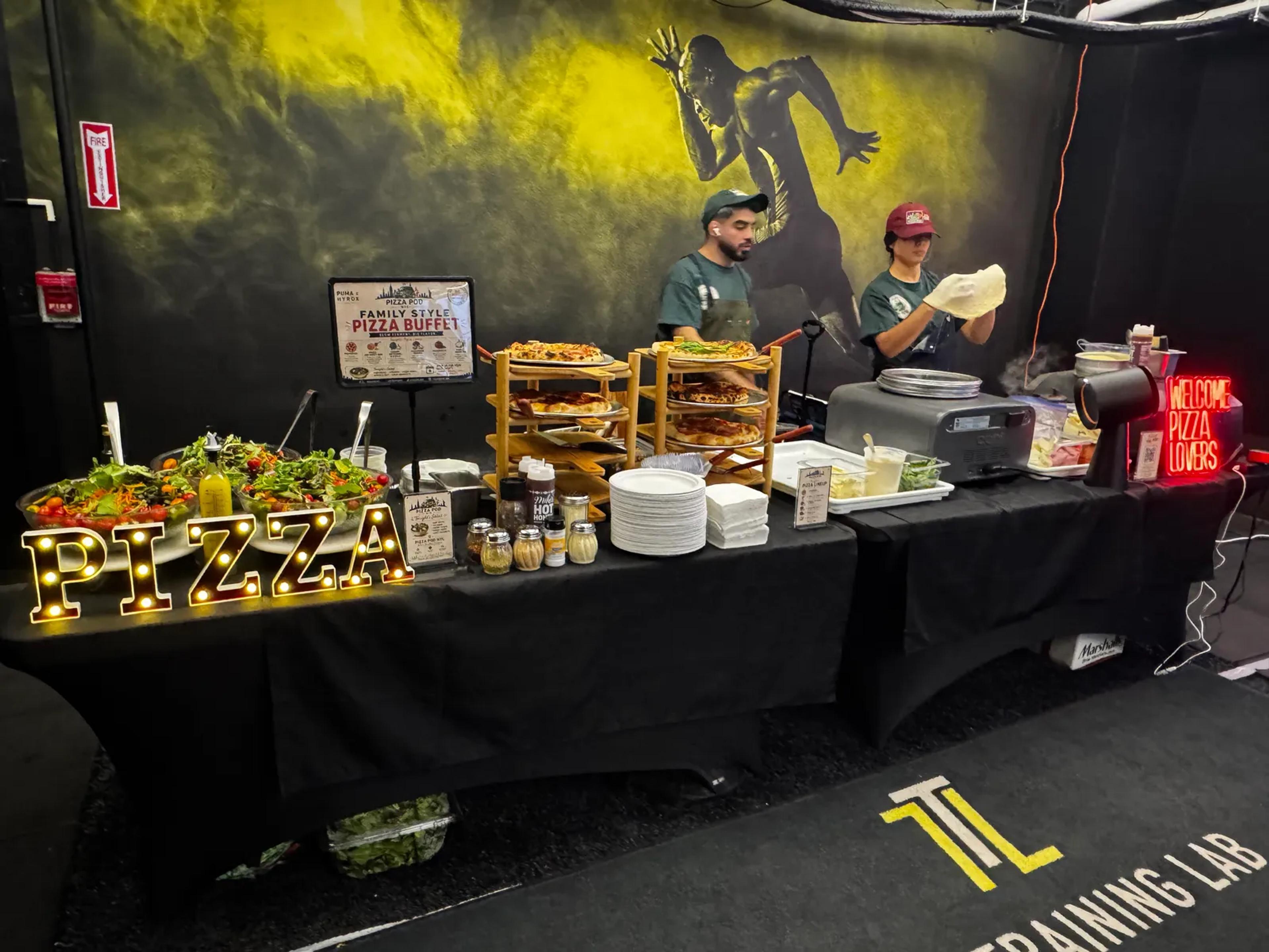 The family-style buffet along black-draped tables against a yellow mural, two cooks behind it and a Training Lab decal on the floor