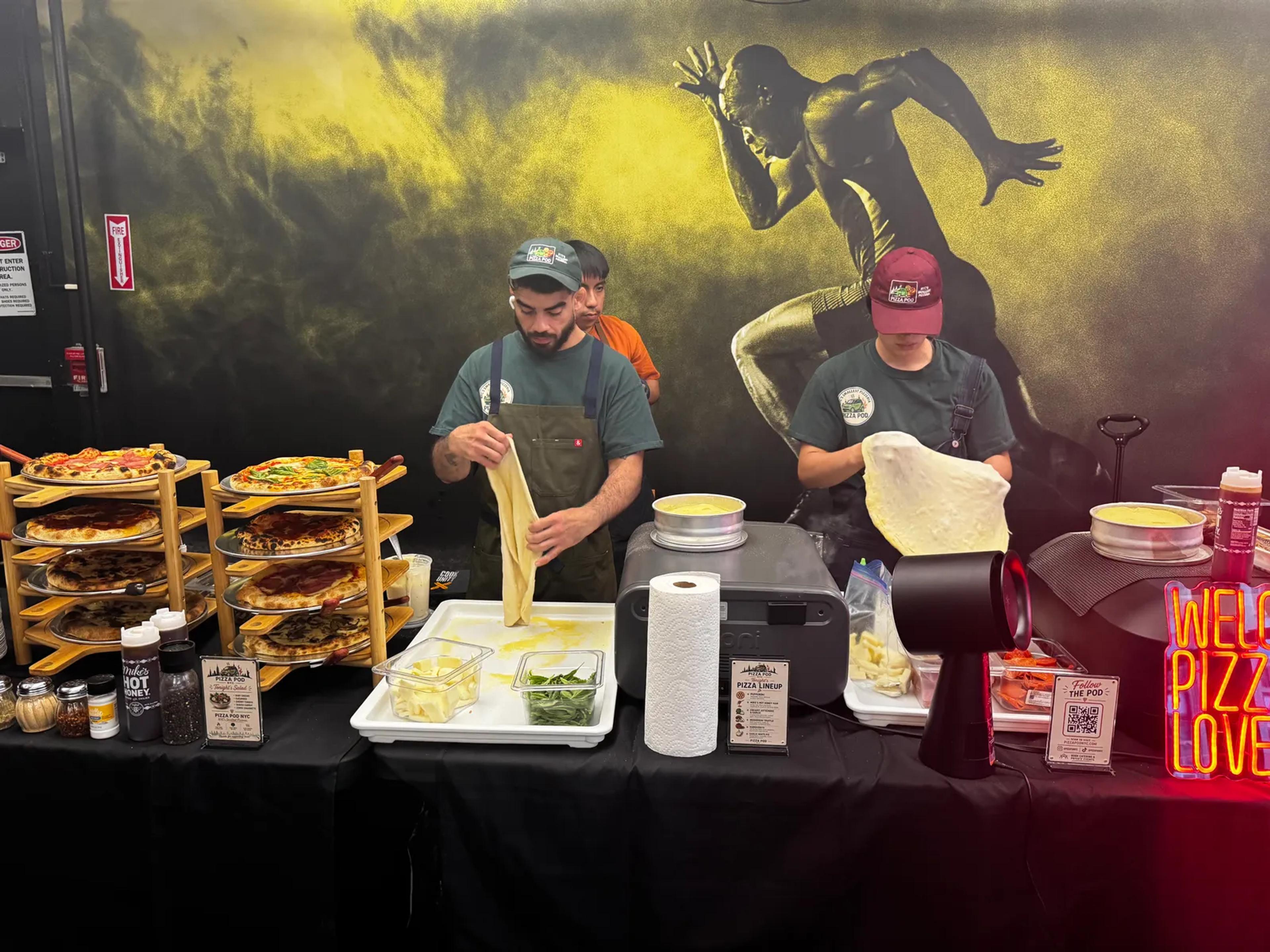 Two cooks stretching dough by hand behind the buffet, racks of finished pies to their left and an oven on the table between them