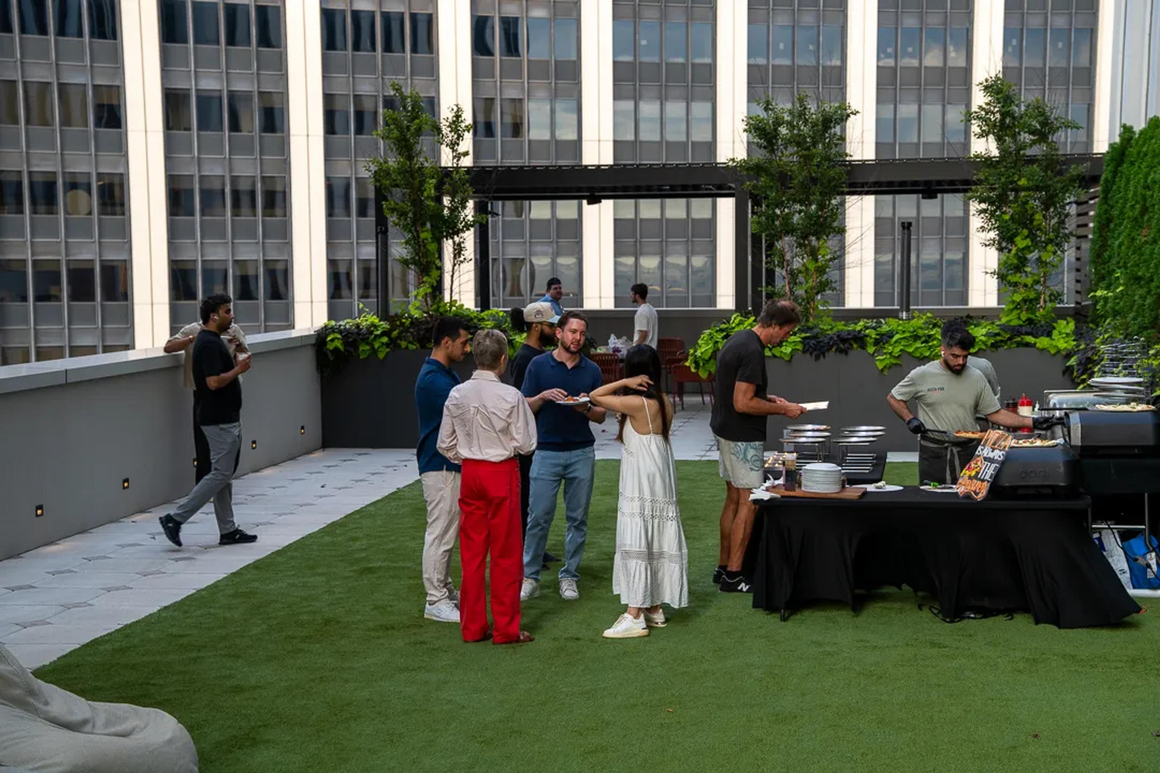 Guests with plates on the rooftop lawn beside the black-draped service table, towers behind them