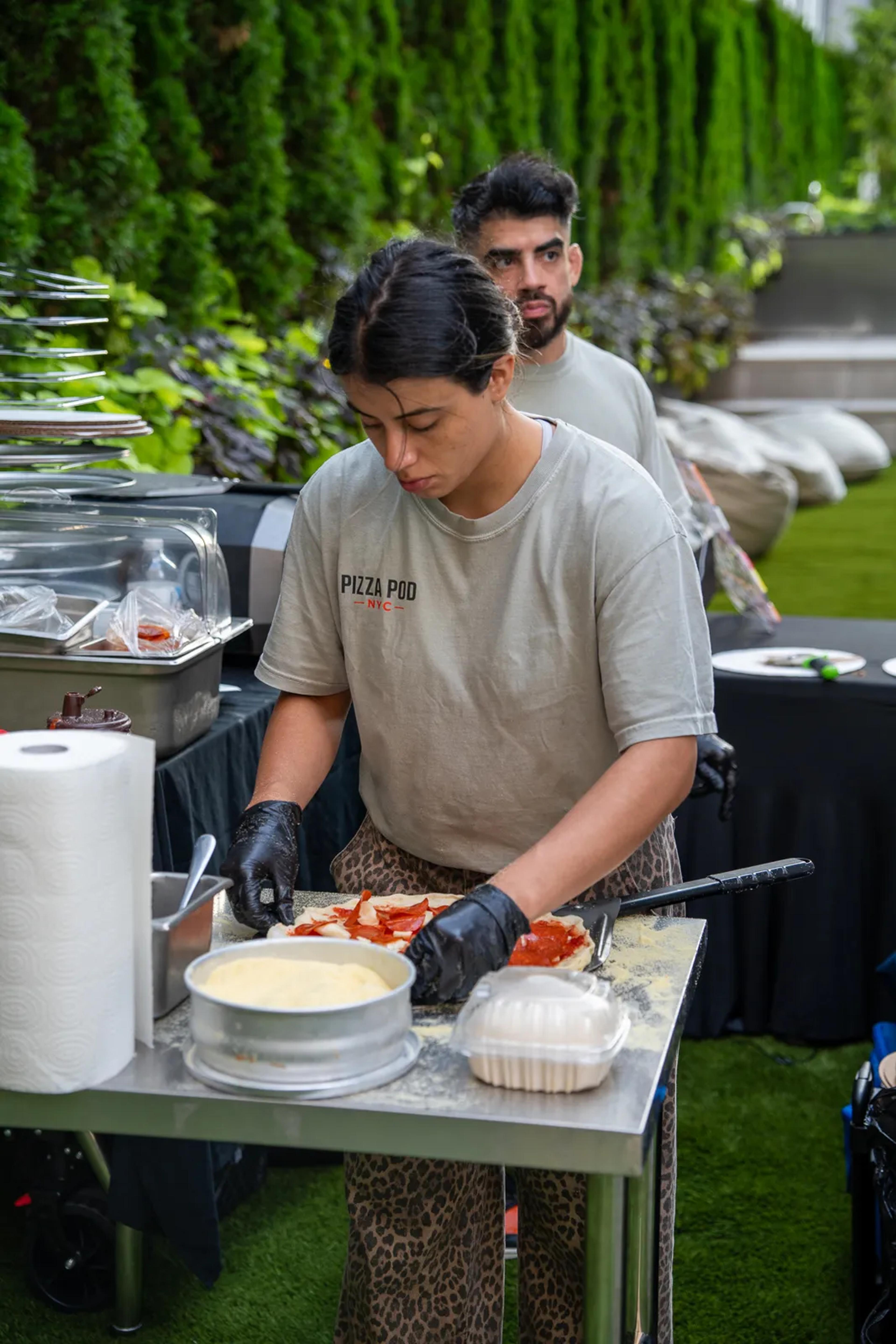A cook in a Pizza Pod shirt topping pies at the prep table on the lawn, a second cook behind her