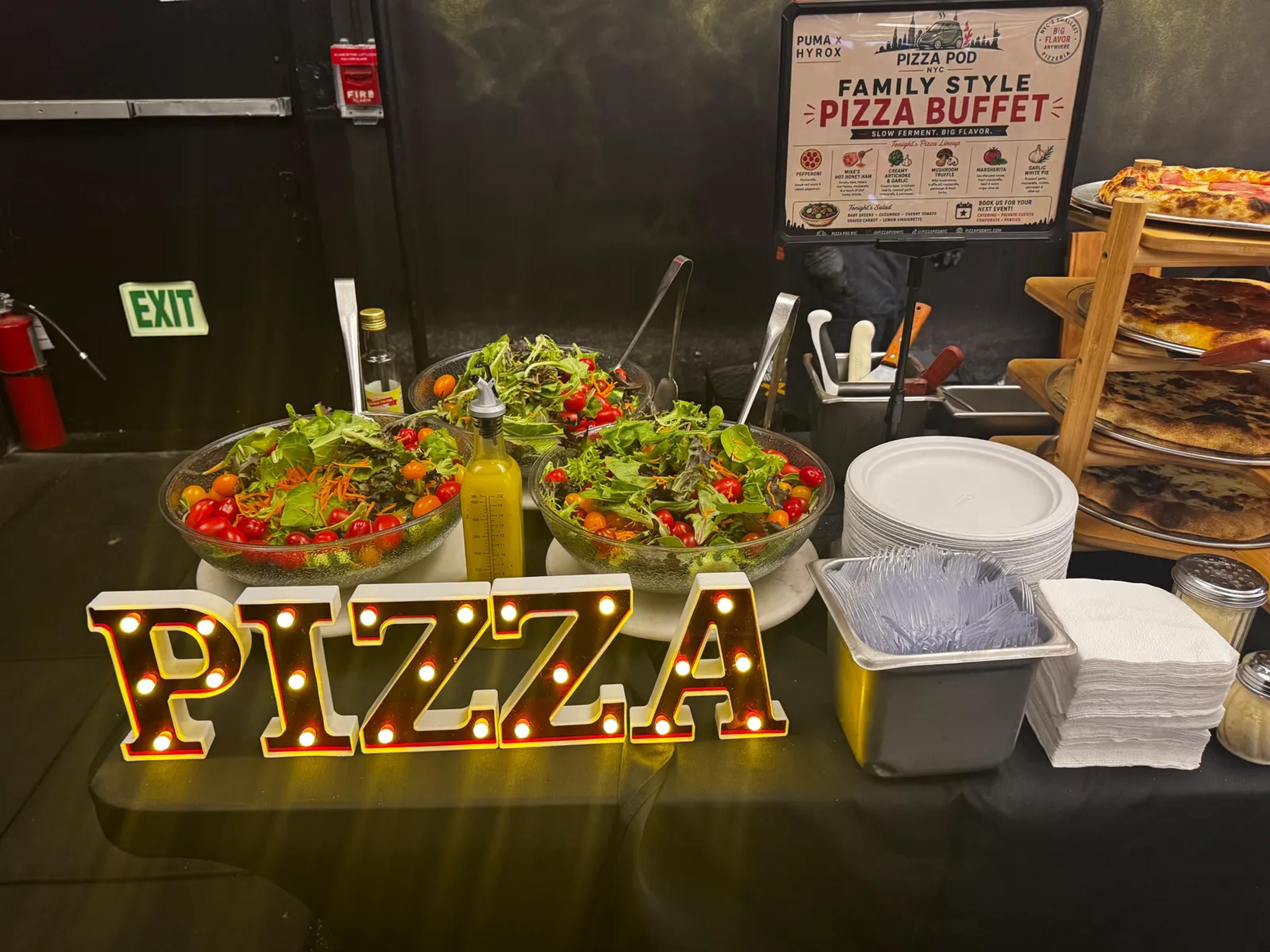 Light-up PIZZA letters in front of bowls of salad, with the PUMA × HYROX family-style pizza buffet board and a rack of pies behind