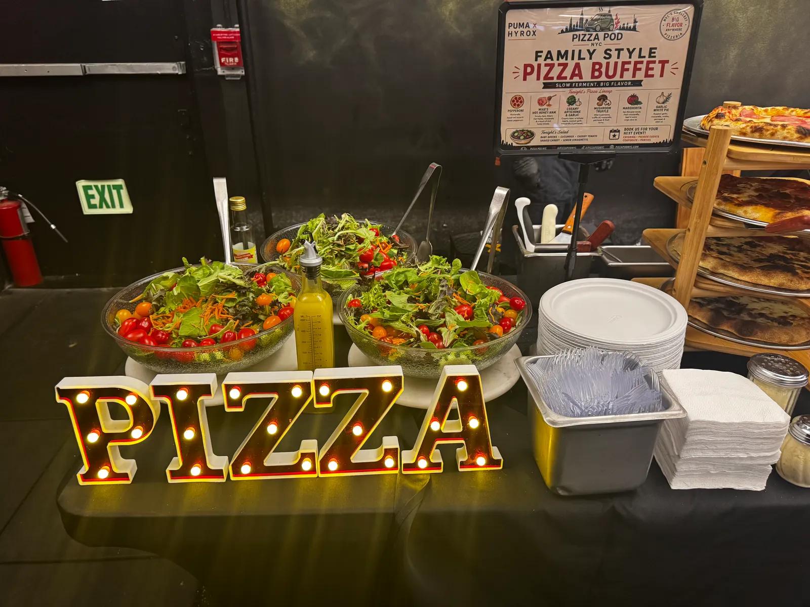 Light-up PIZZA letters in front of bowls of salad, with the PUMA × HYROX family-style pizza buffet board and a rack of pies behind