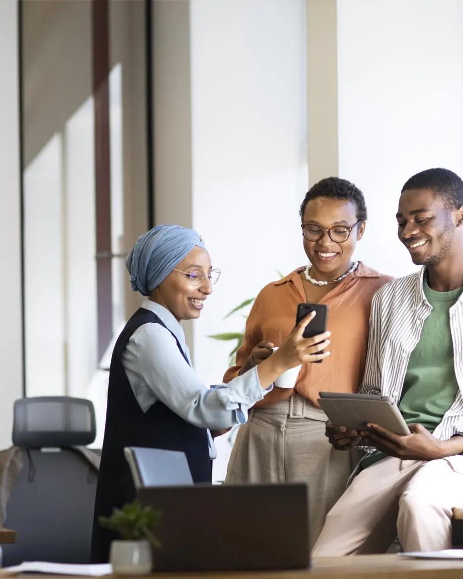 Trois jeunes professionnels, dont une femme voilée, regardent ensemble l'écran d'un téléphone dans un bureau.
