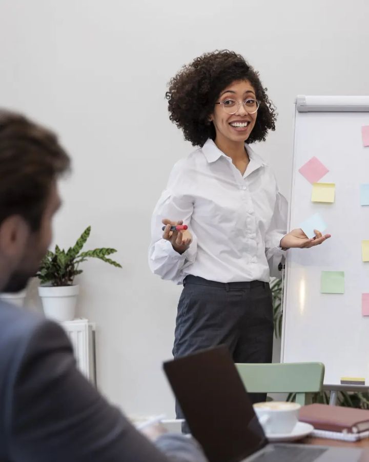 Une femme présente un tableau couvert de notes adhésives à des collègues attablés.
