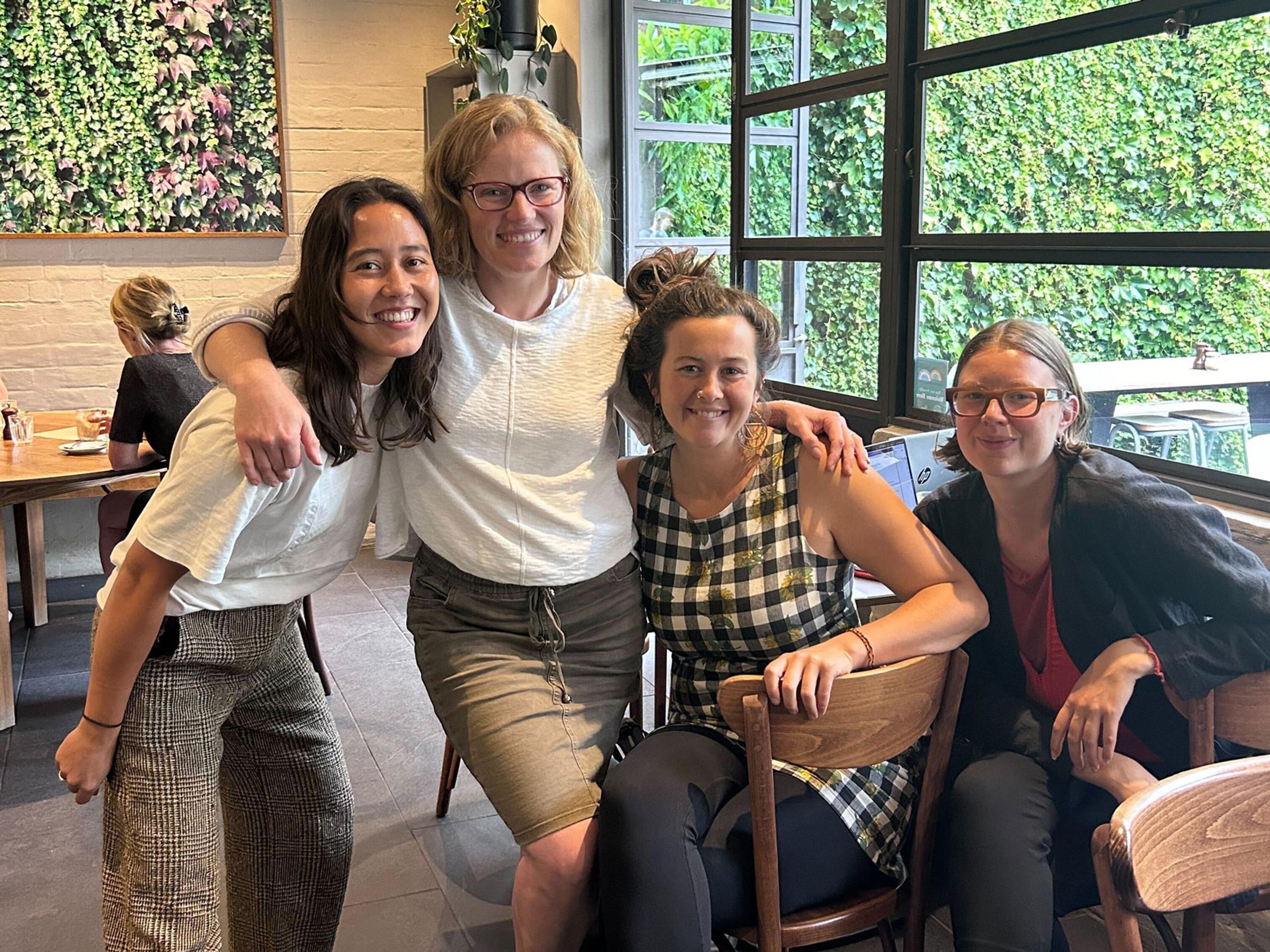 Four women sitting and standing in a cafe space smiling at the camera.