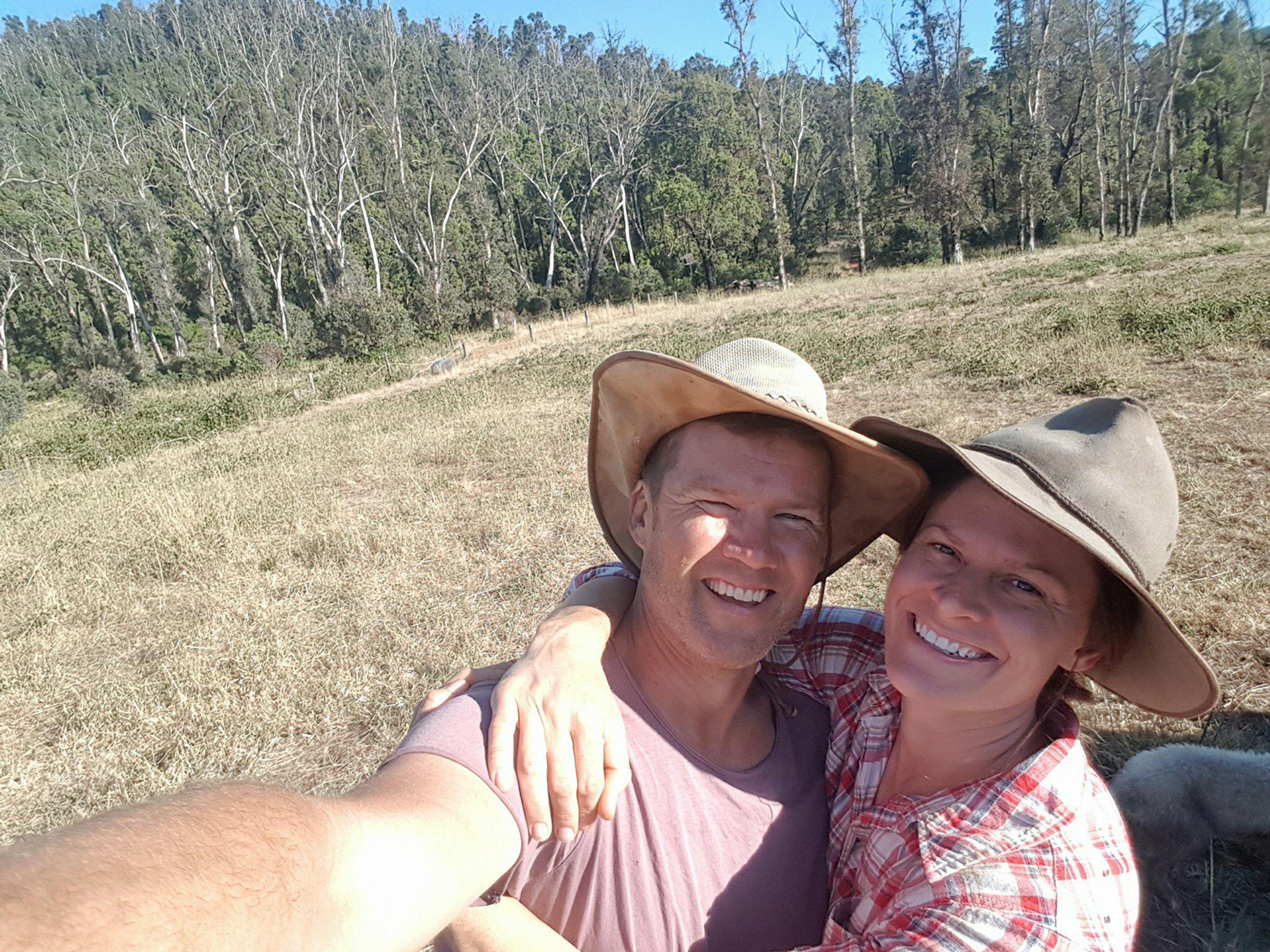 Two people taking a selfie in a paddock on a sunny day. The sky is blue and there are a lot of trees in the background next to the paddock. The people are smiling and hugging.
