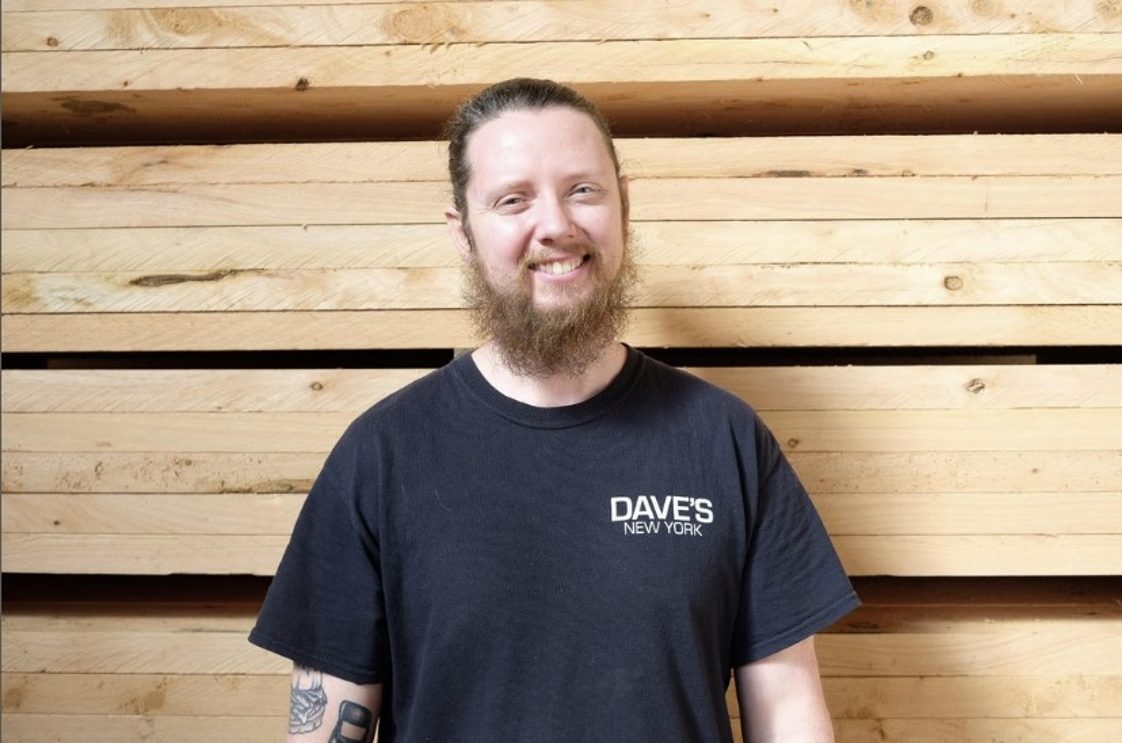 A person in a black t-shirt smiling at the camera in front of a stack of wooden planks.