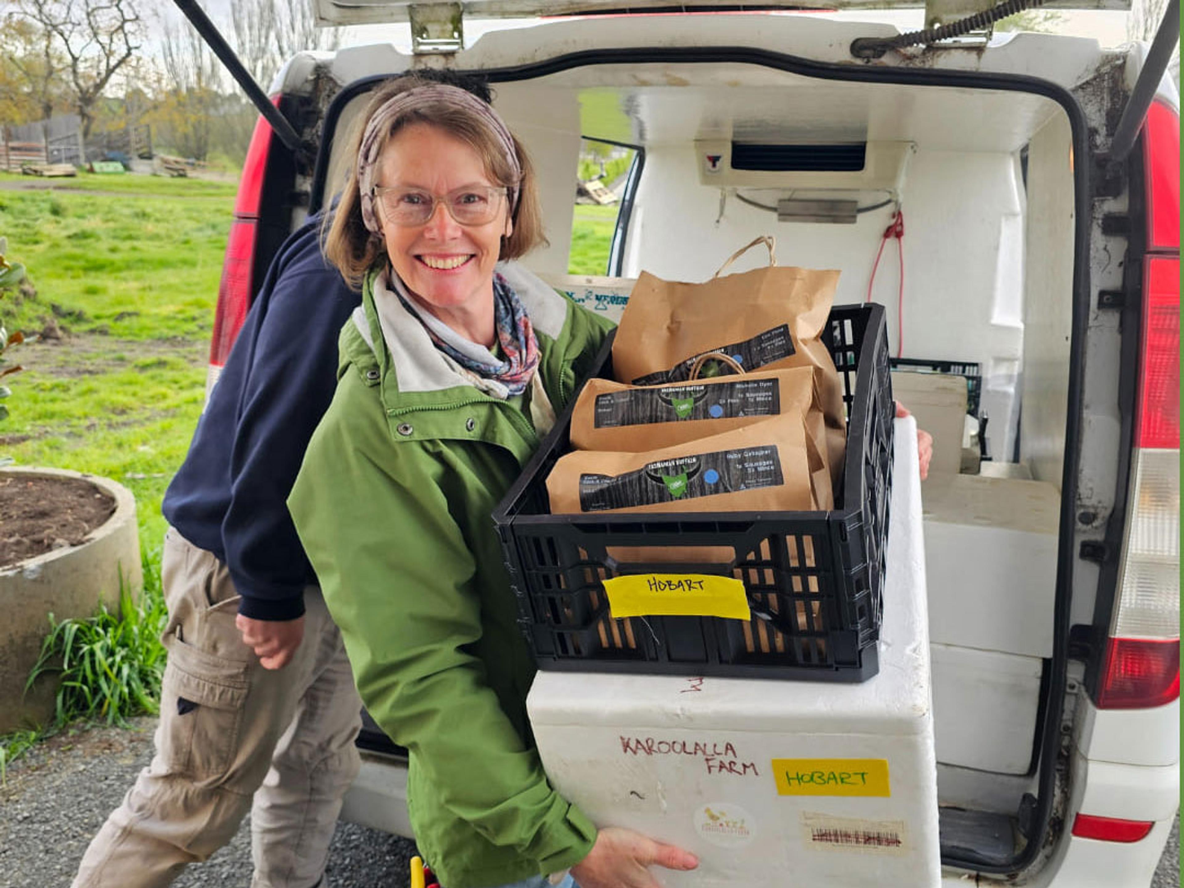 A smiling woman loading a crate and a cool box into a van. The crate has paper bags presumably filled with fresh food.