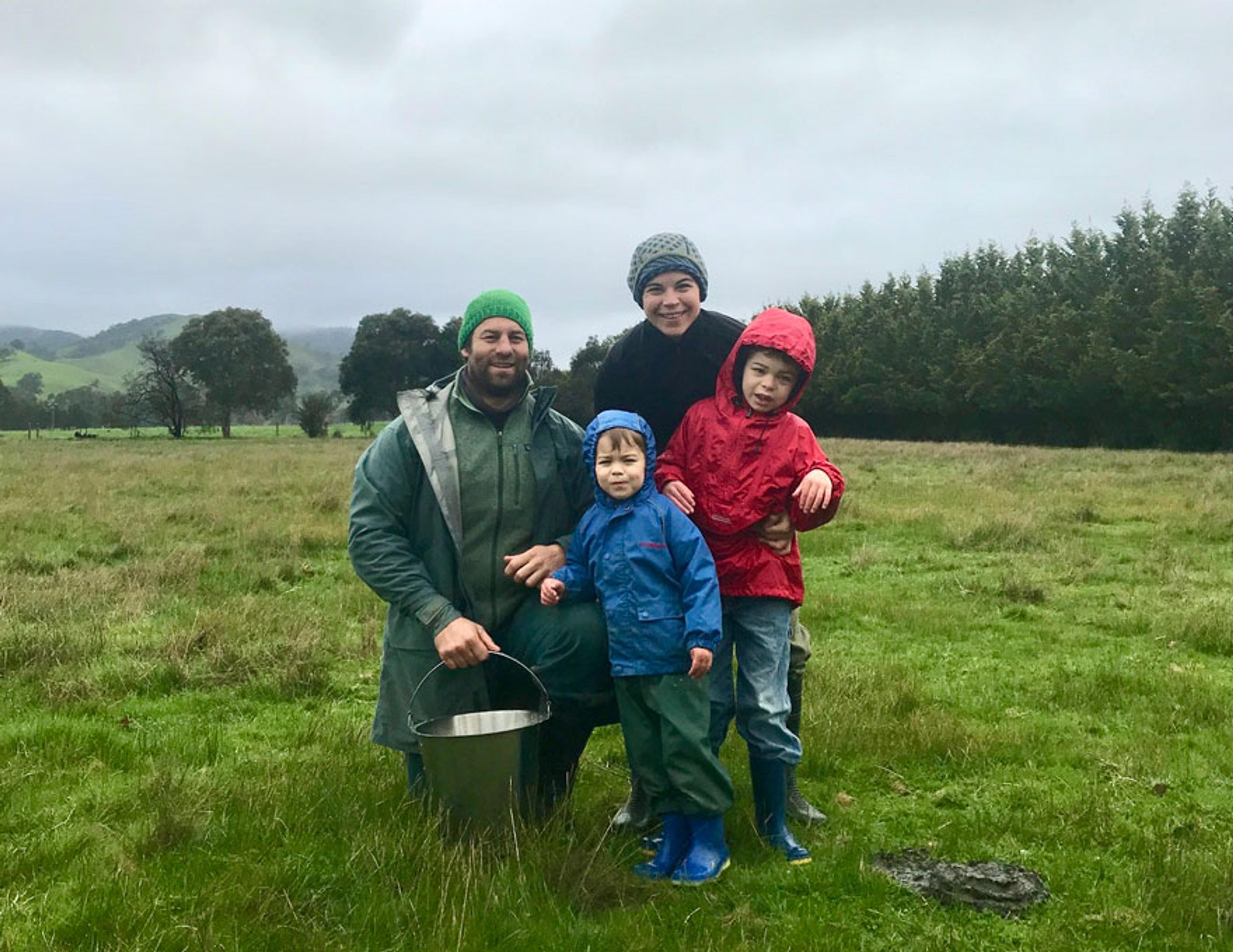 A couple and their two children posing for the camera. They're all rugged up with beanies and jackets. They're in the middle of a paddock with grey skies and a row of trees behind them. 