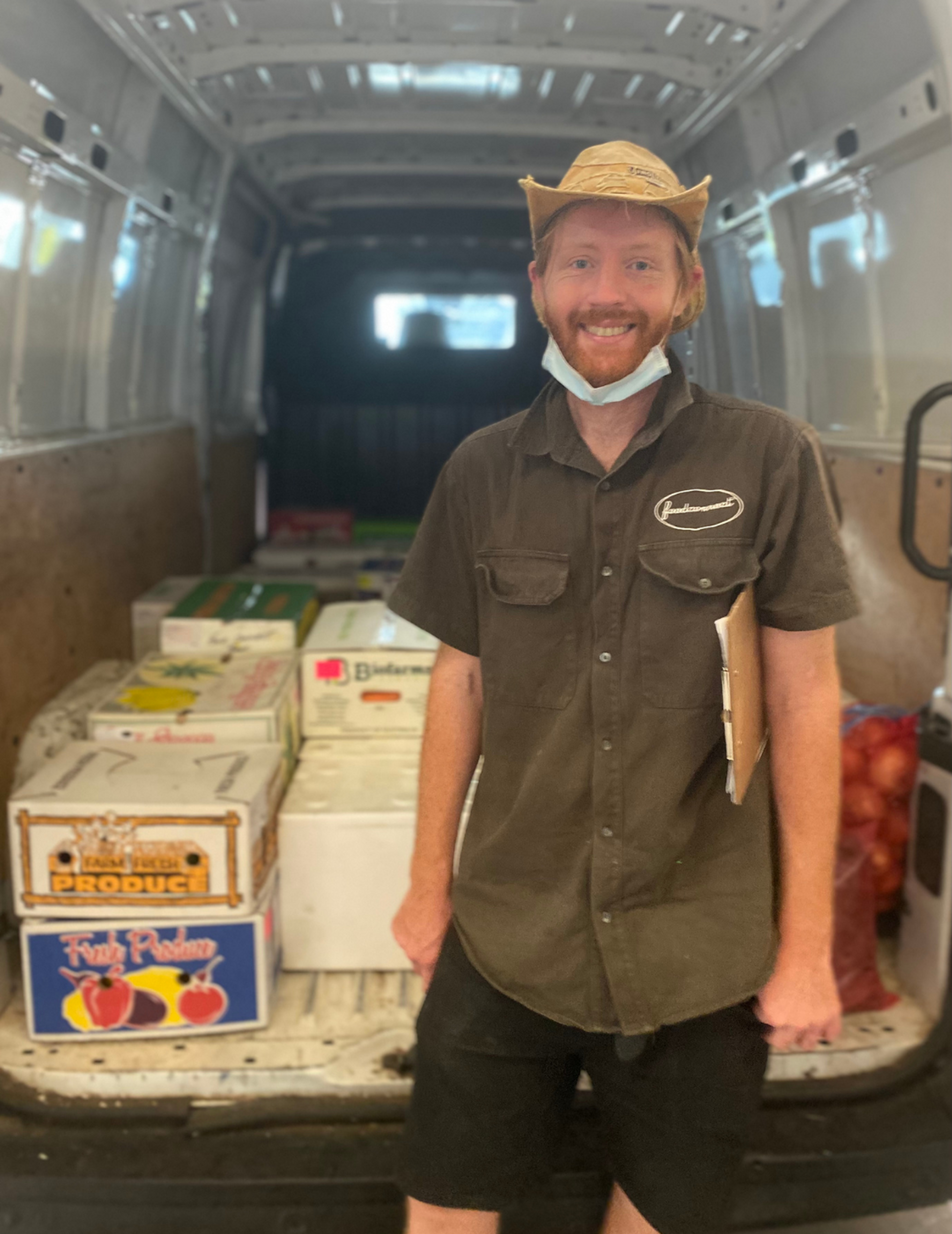 A person standing in front of the back of a van that contains some boxes of produce. They are smiling.