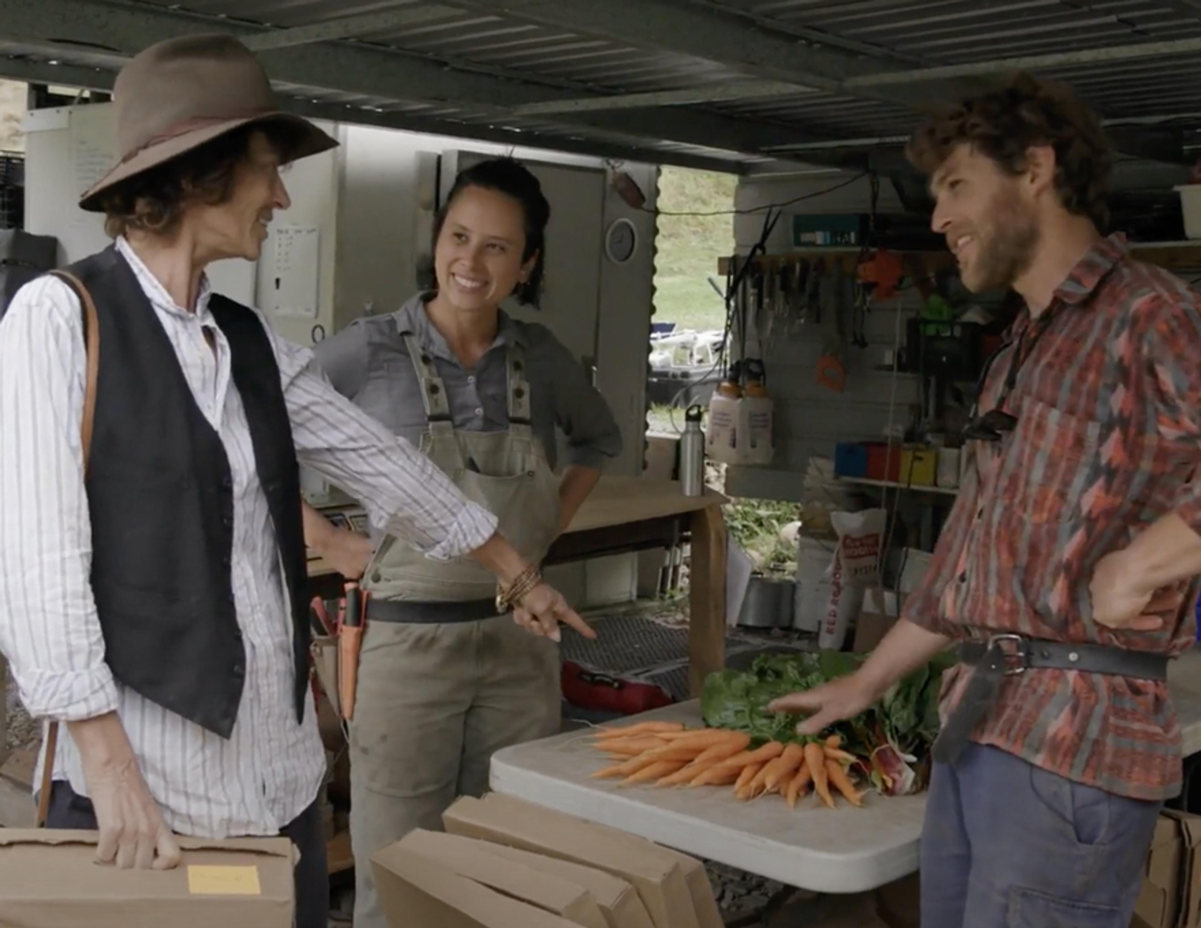 People standing around chatting in an open shed with a bunch of carrots on the table.