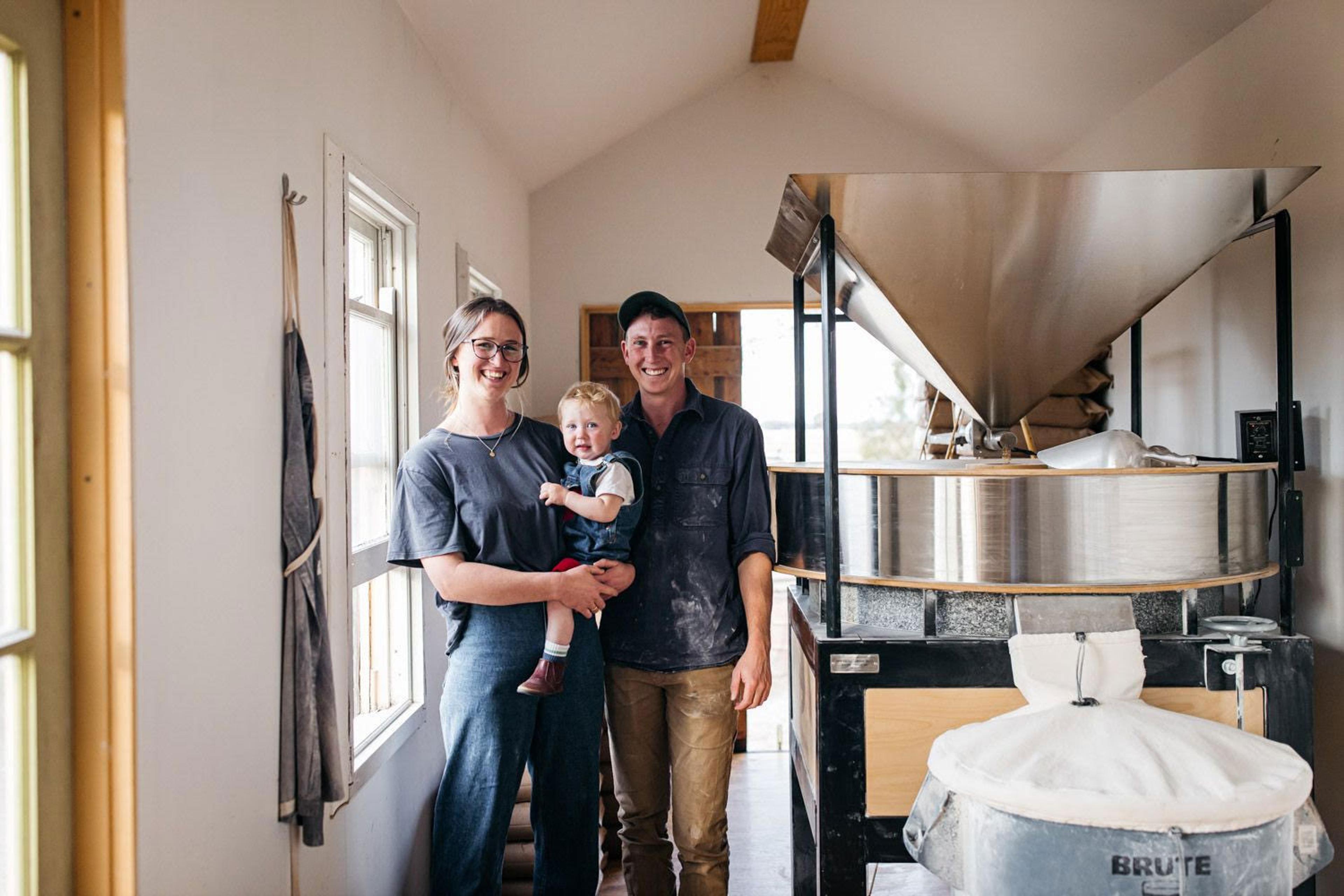 Courtney and Ian with their daughter standing next to a stone mill. They are all smiling at the camera.