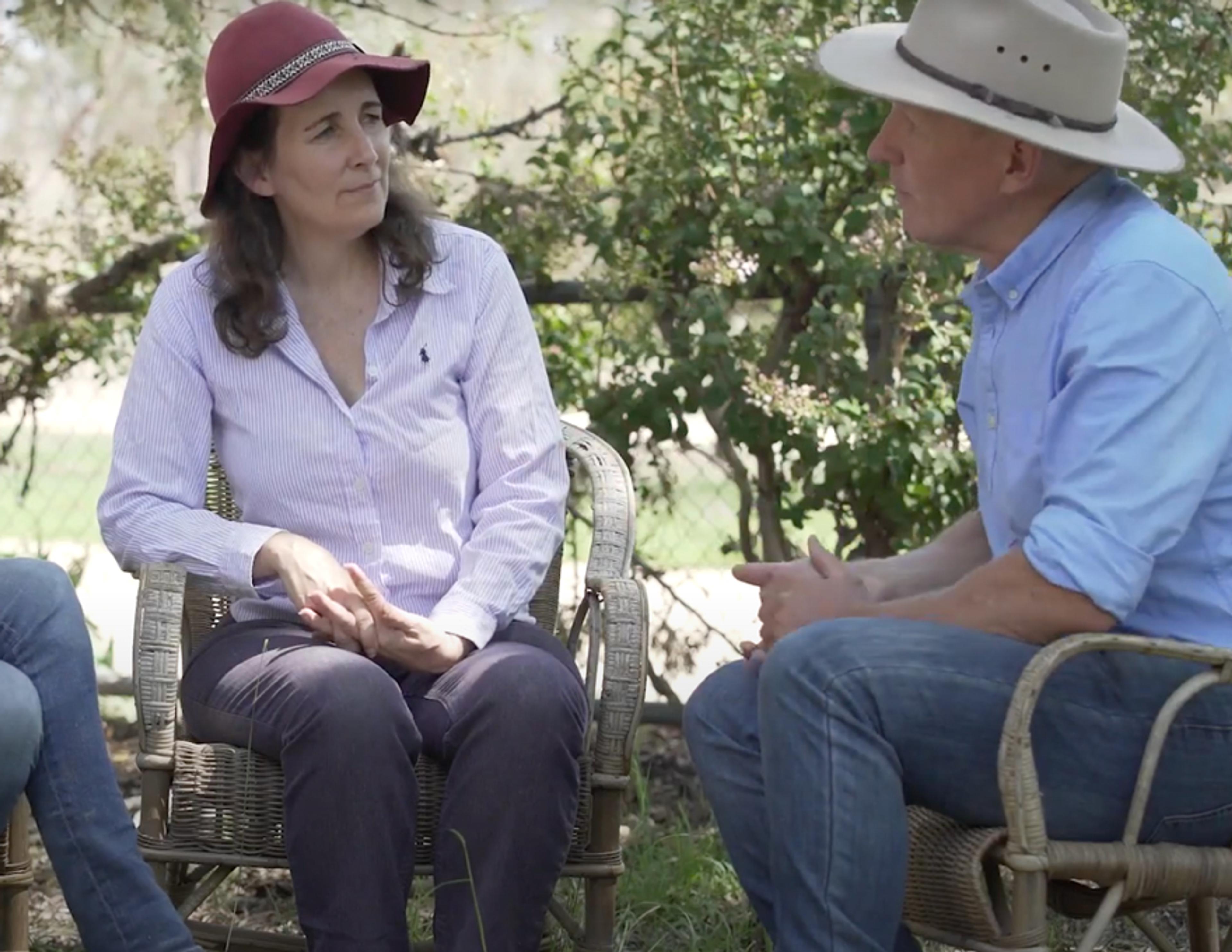A husband and wife farming team sitting on chairs outside with hats on, discussing regenerative agriculture.