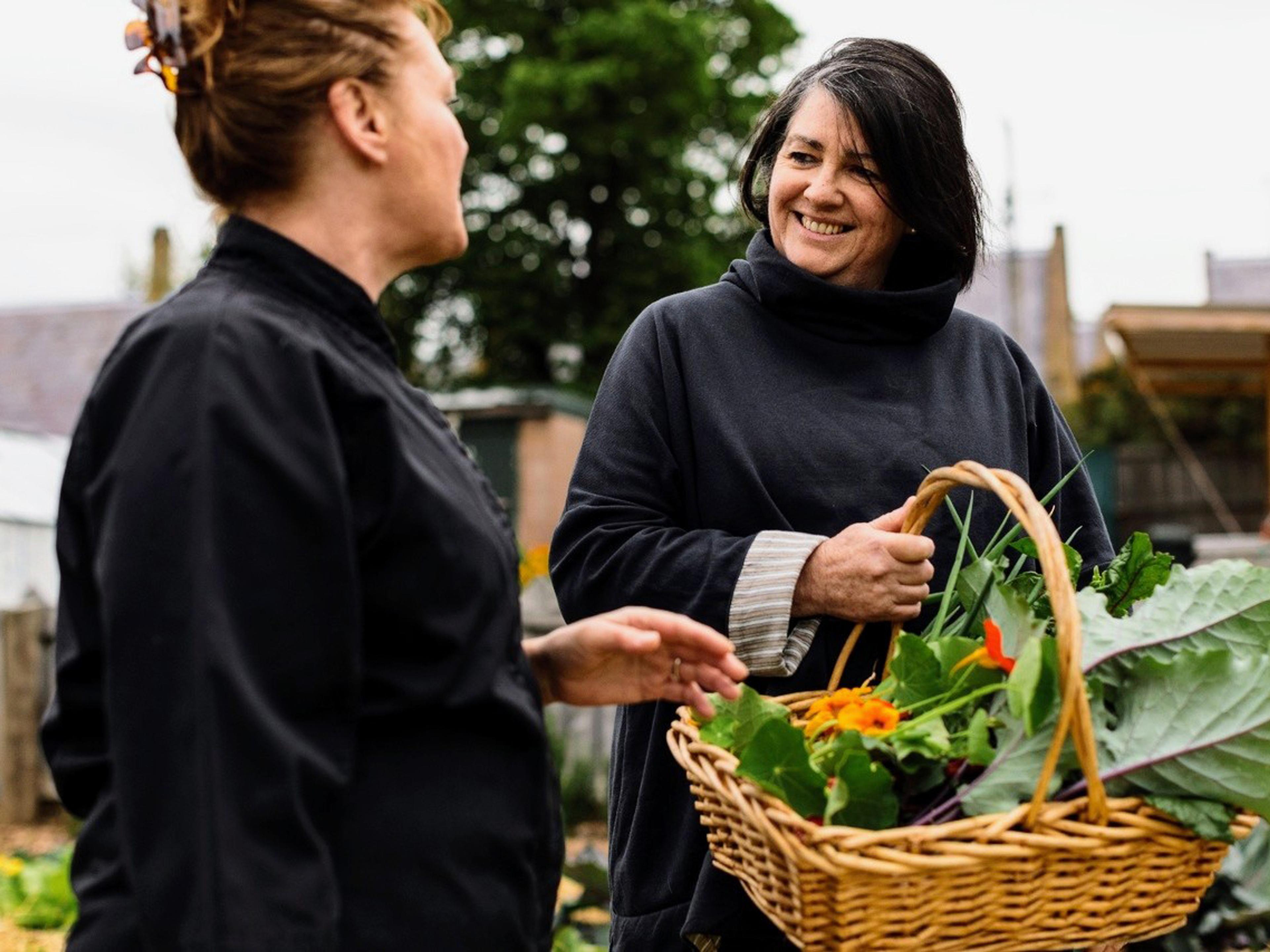 A person holding a basket of fresh produce smiling at another person who is dressed as if they work in hospitality. They are outside and there is a tree in the background.