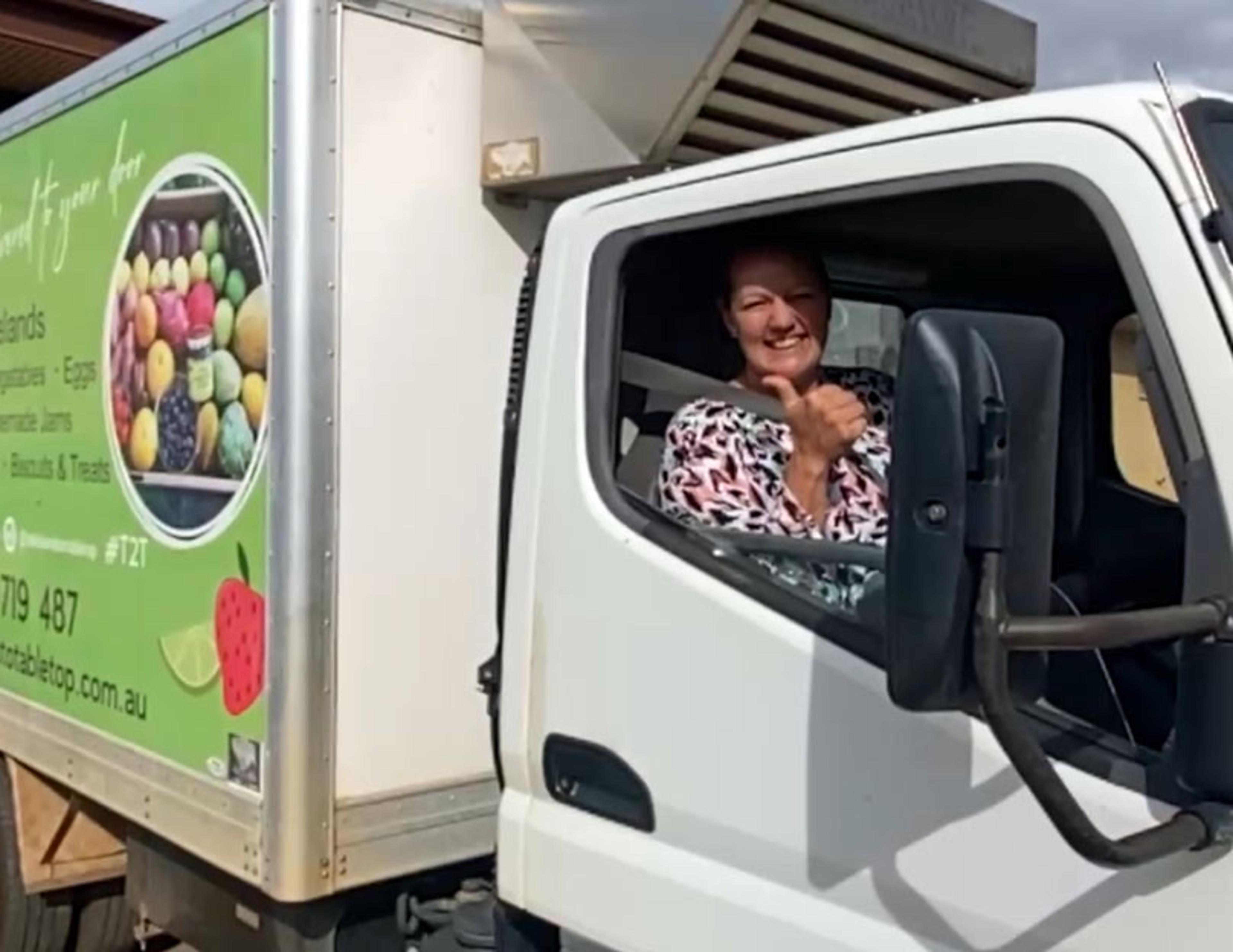 A smiling woman sitting behind the wheel of a small delivery truck. She is giving the camera a big thumbs up.