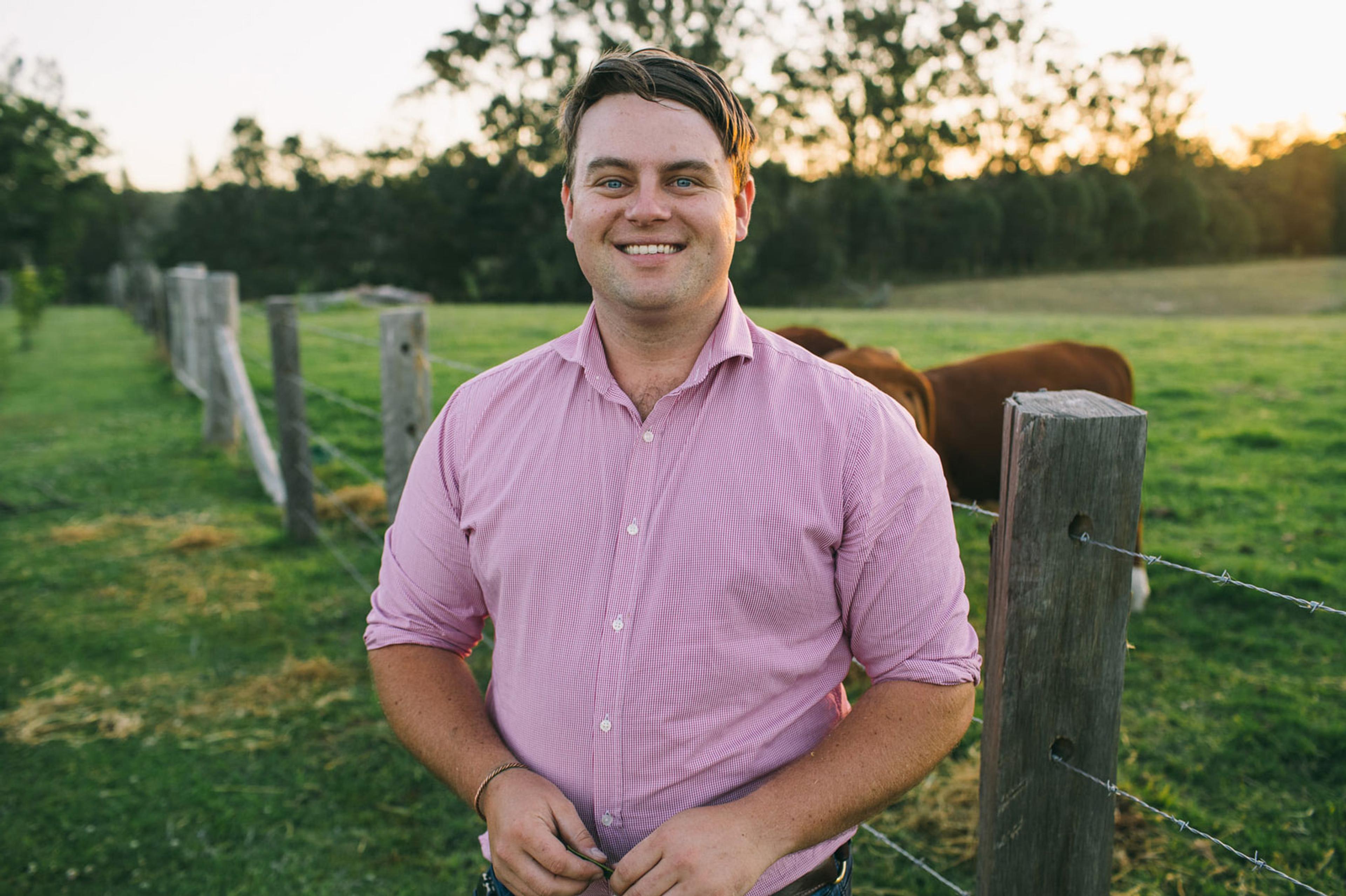 A man in a pink striped short sleeved shirt smiling at the camera. He is standing in a paddock near a wire fence. There are brown cows behind him.