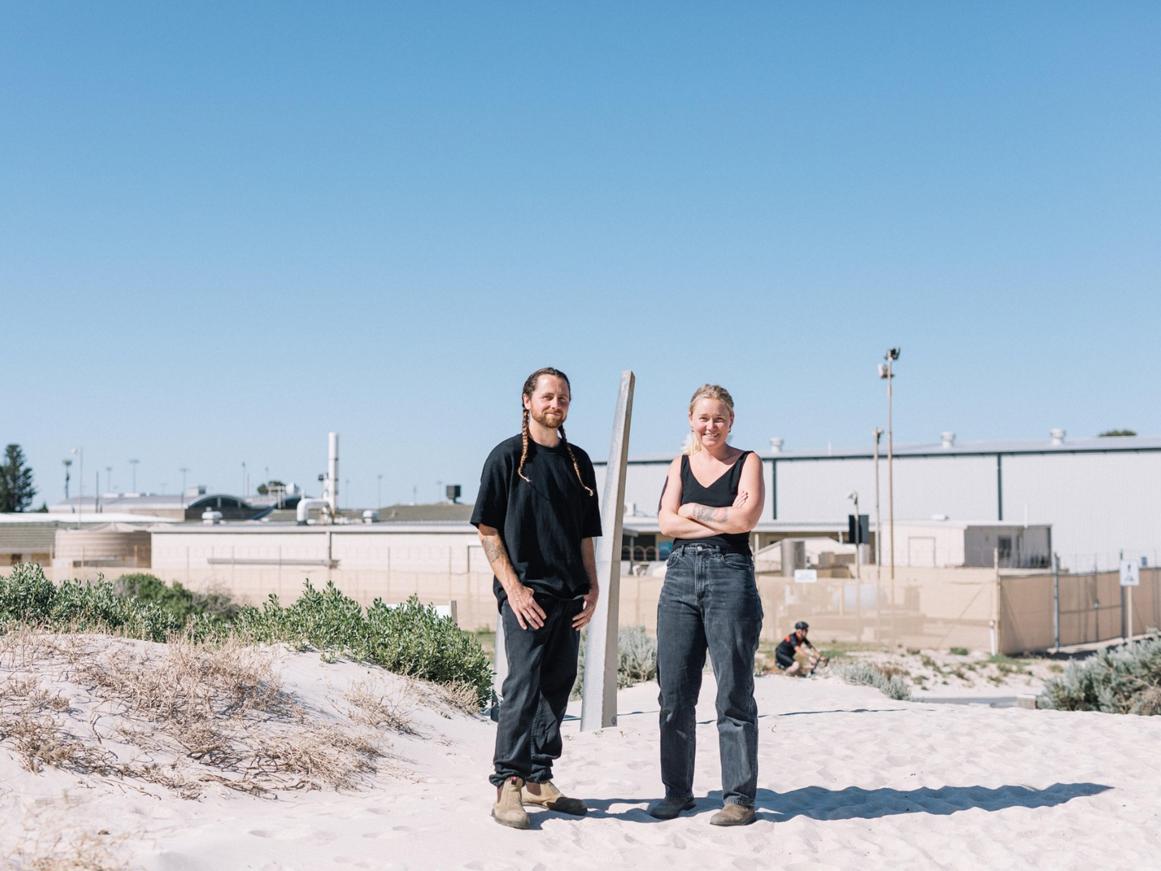 Two people dressed in black at the beach looking directly at the camera.
