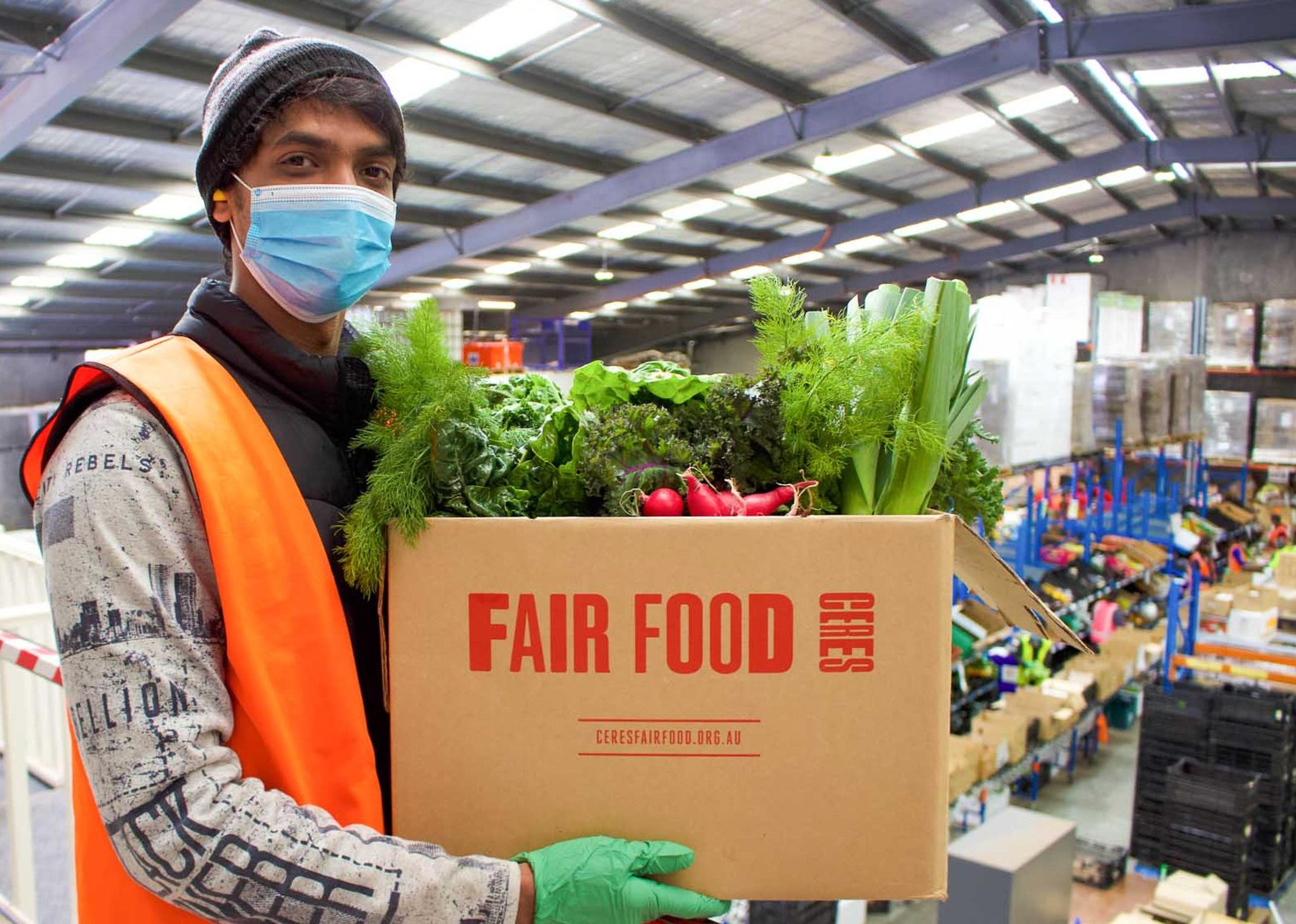 A person wearing a mask and a beanie, standing in a warehouse, holding a cardboard box with the words 'Fair Food CERES' on it. Inside the box is lots of fresh veggies.