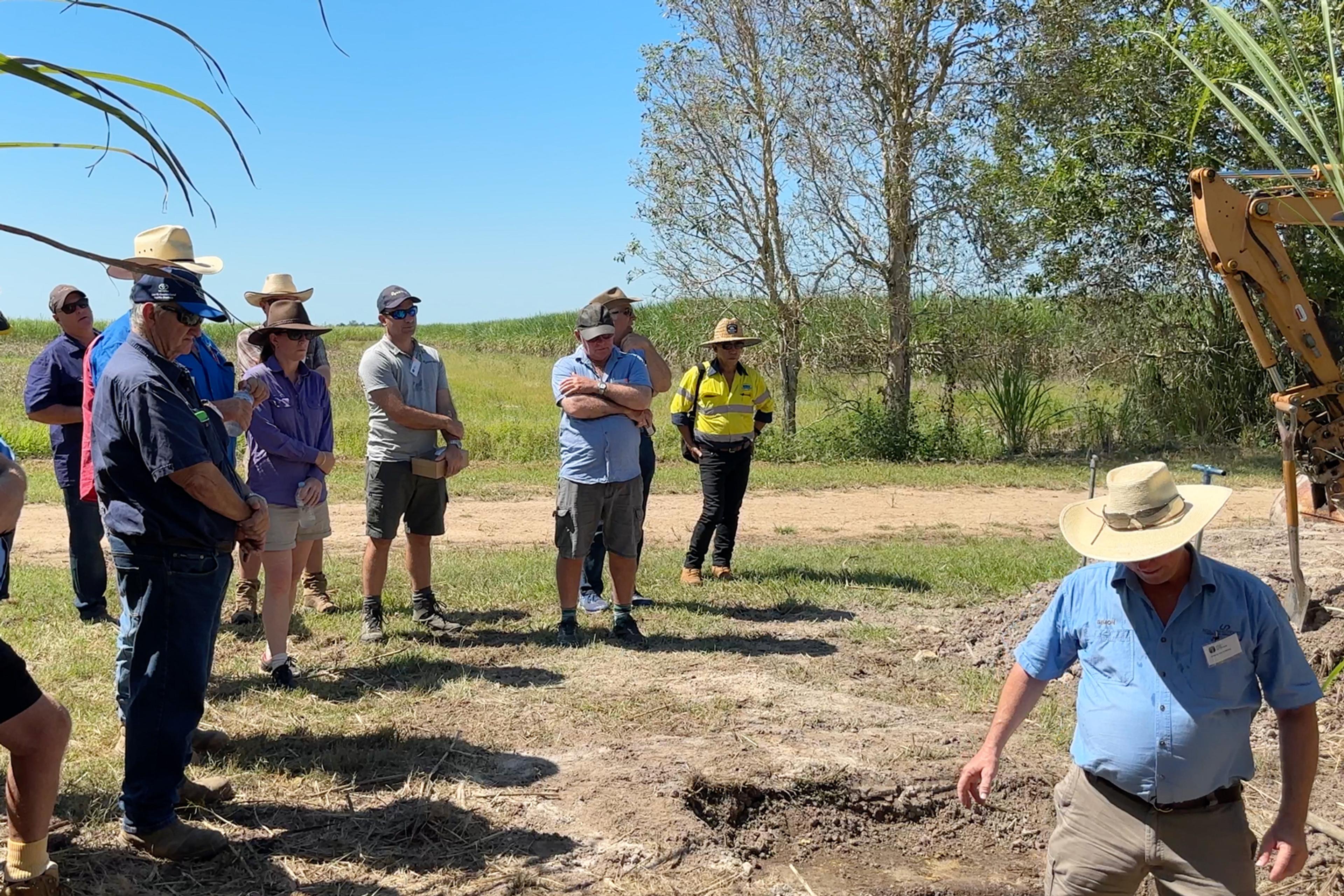A group of people outside watching a man presenting a workshop in a paddock. The skies are blue.