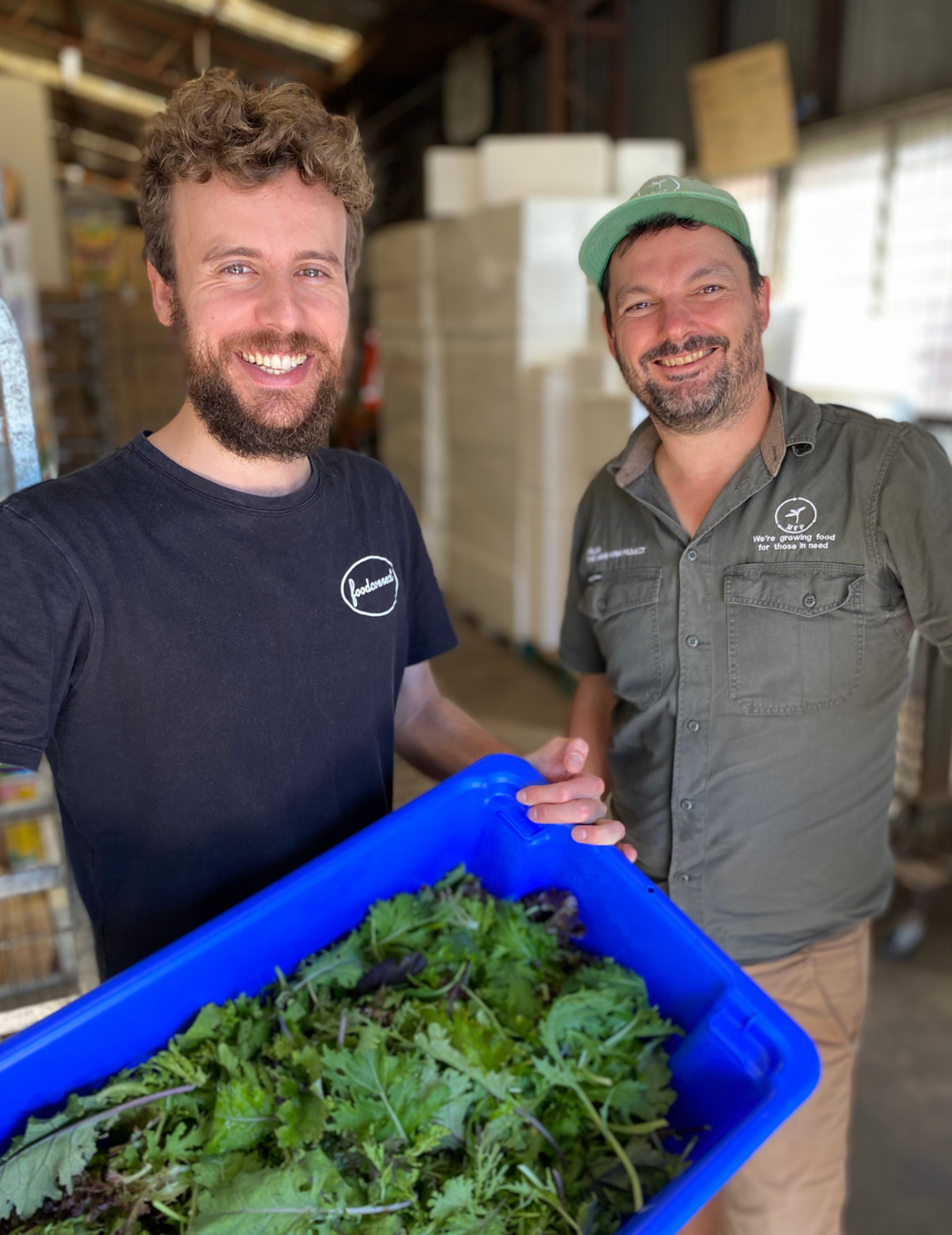 Two people smiling at the camera. One of them is holding a blue box full of greens.