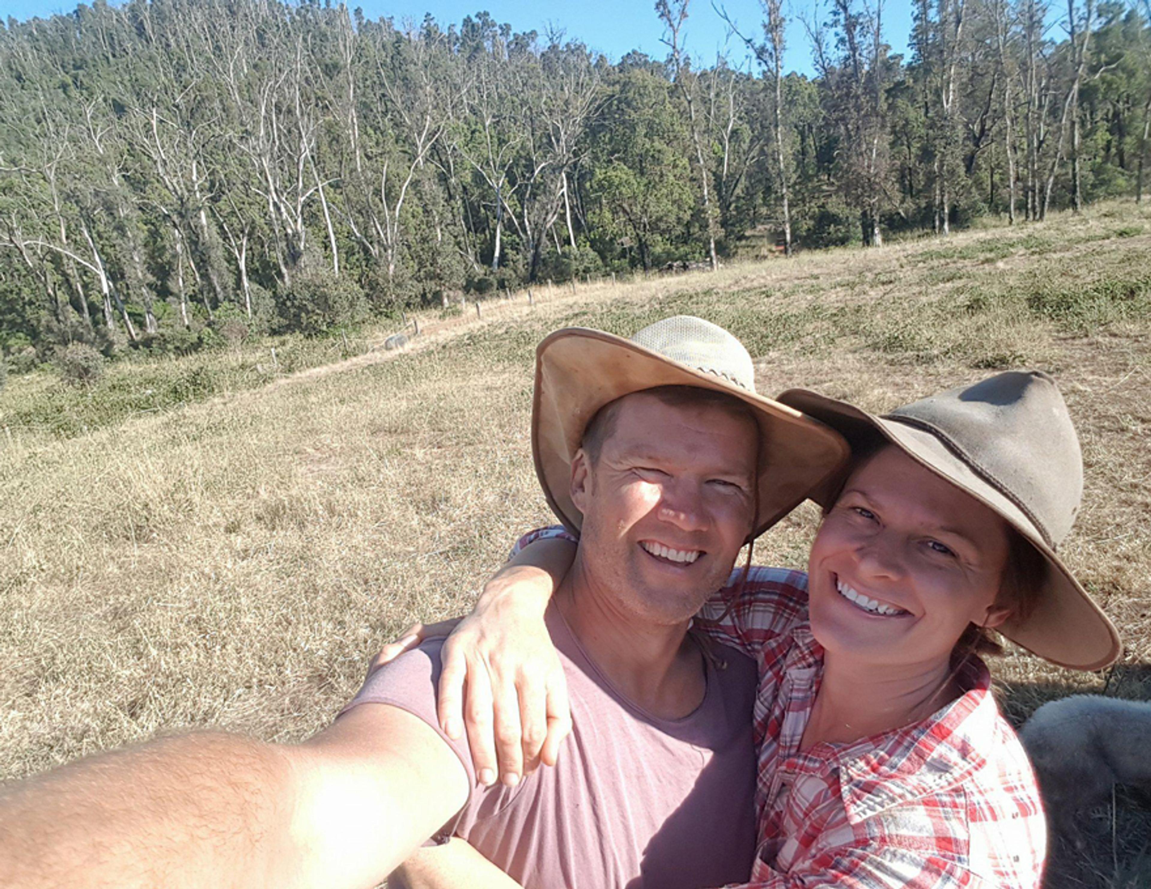 Two people taking a selfie in a paddock on a sunny day. The sky is blue and there are a lot of trees in the background next to the paddock. The people are smiling and hugging.
