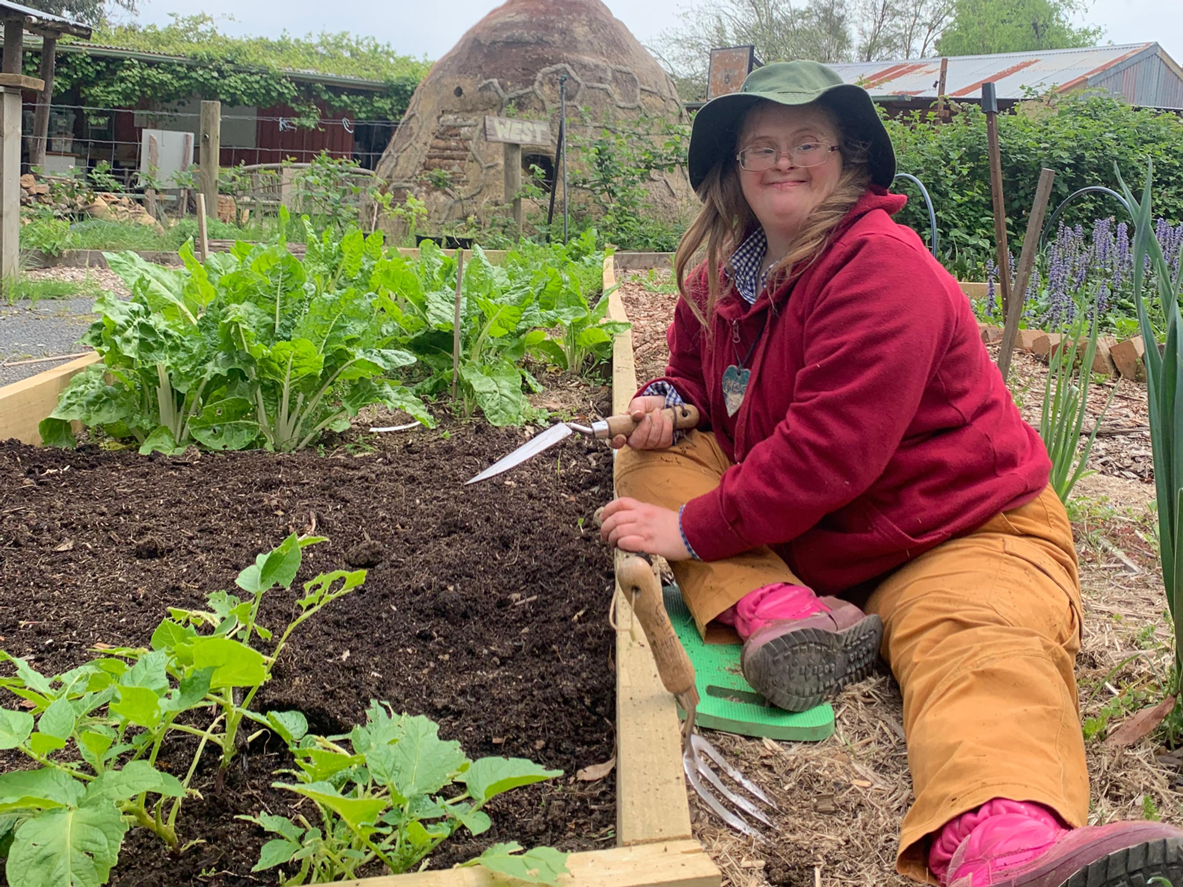 A person sitting on the ground next to a garden bed. They are smiling at the camera and holding a trowel over the soil.