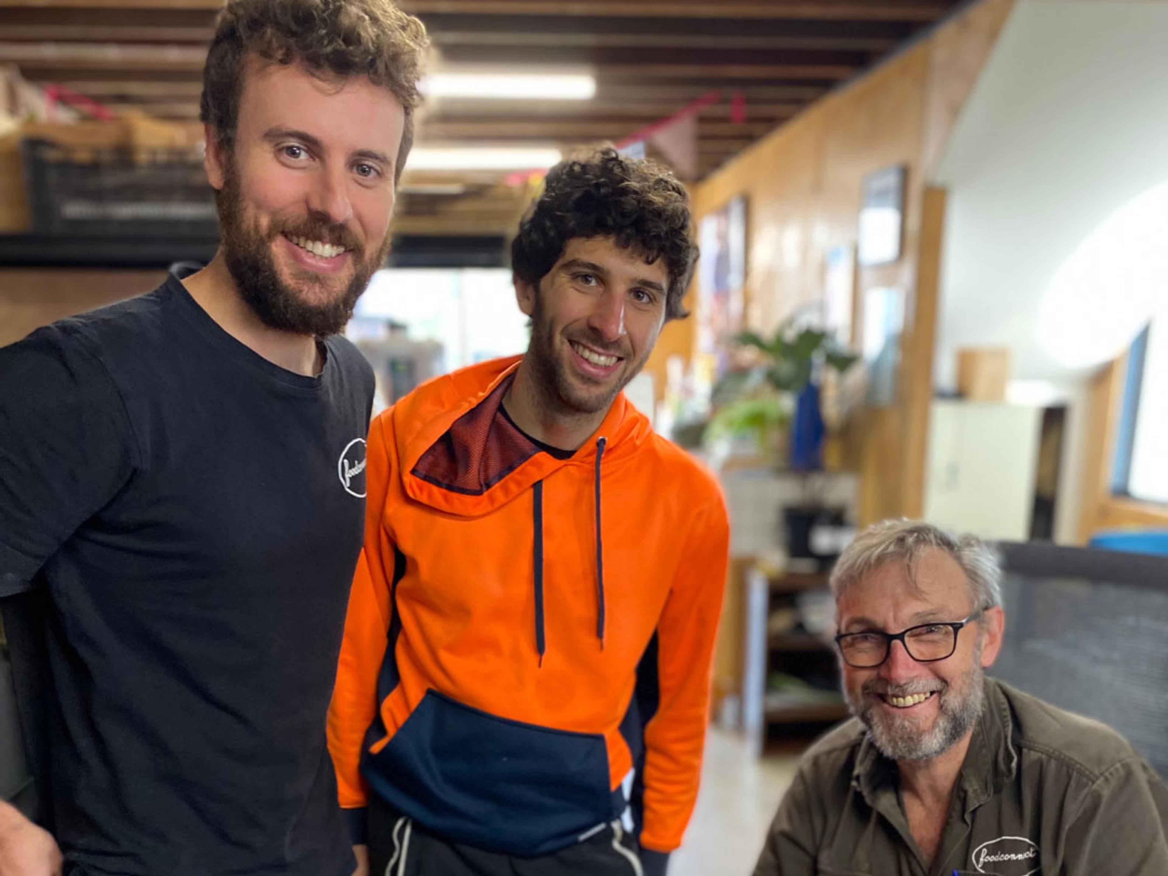 Two people standing and one person sitting. All of them are smiling at the camera.