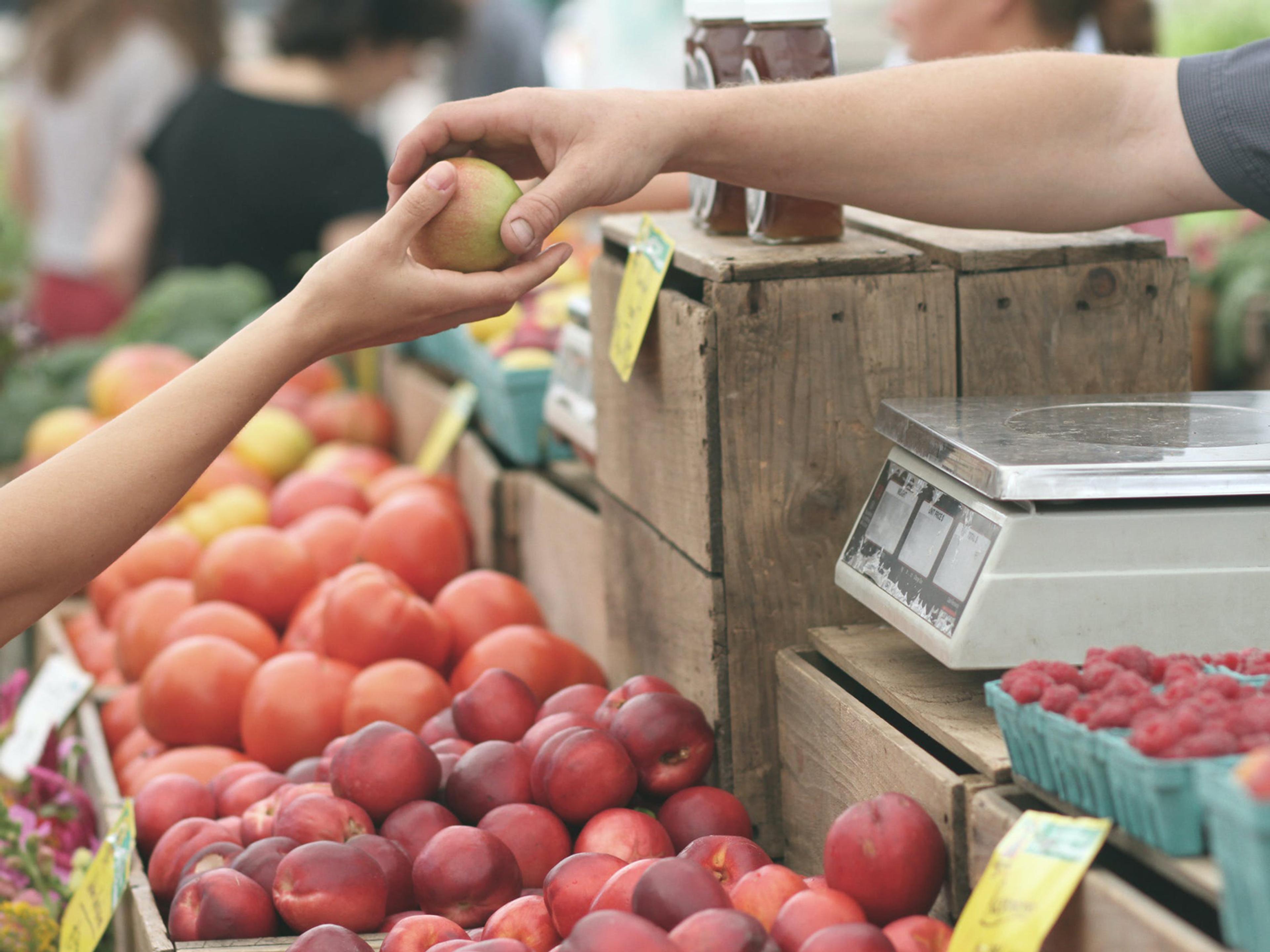 A hand reaching out to take an apple from another hand at a farmer's market.