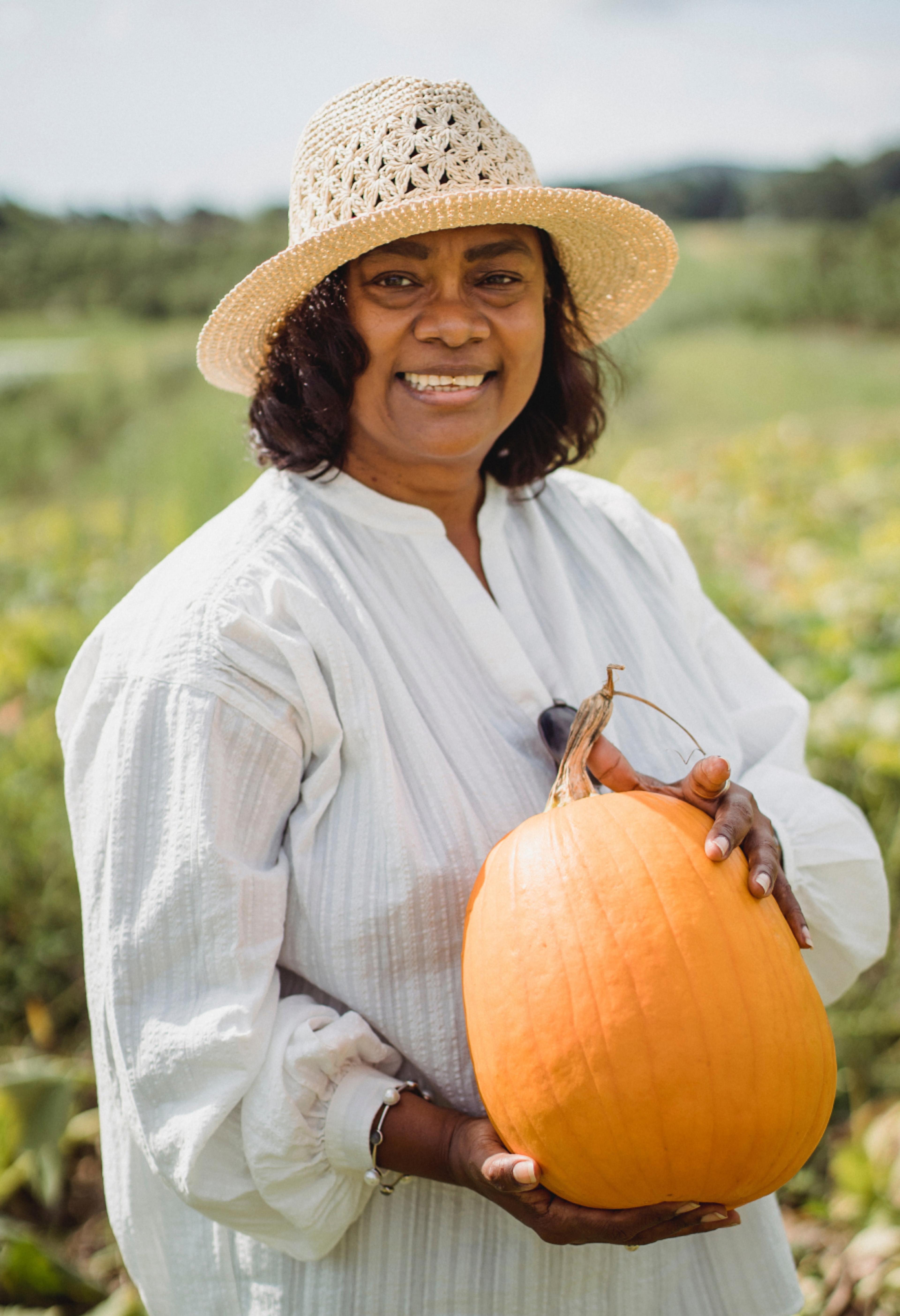 A person with a hat in a field, holding a very large pumpkin.