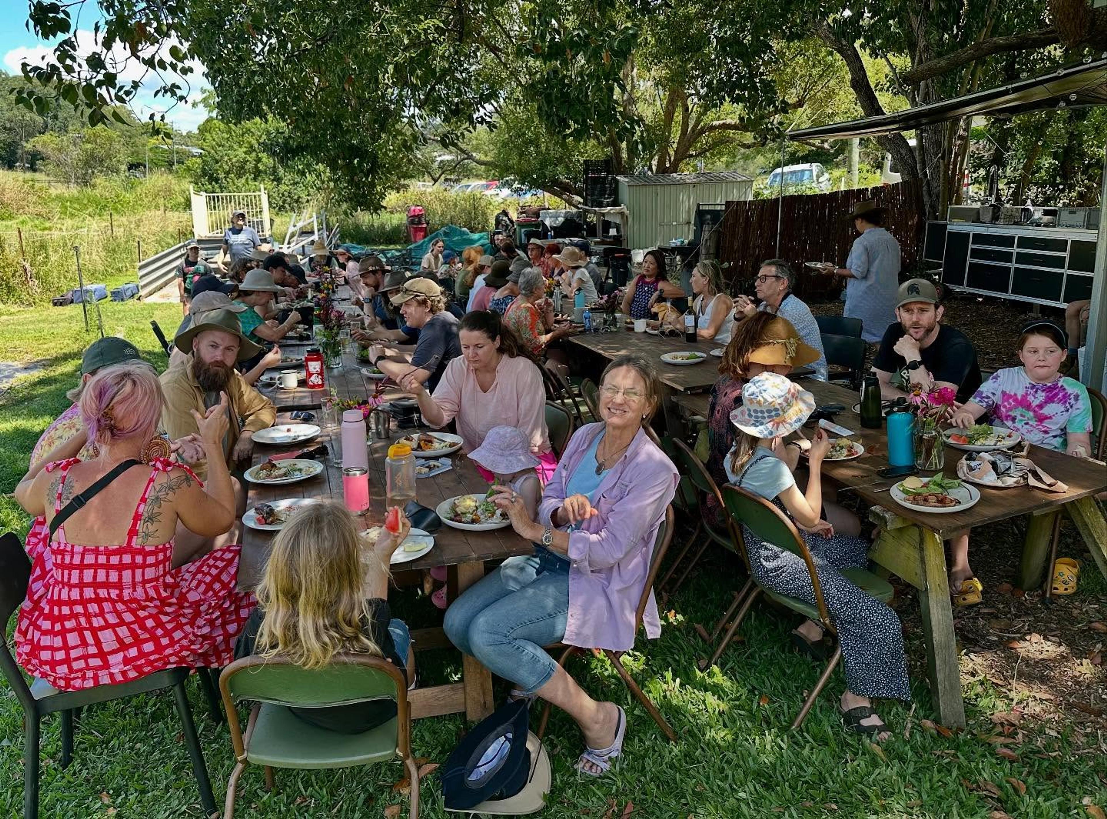 Two long tables filled with happy people eating fresh food under the shade of a big tree.