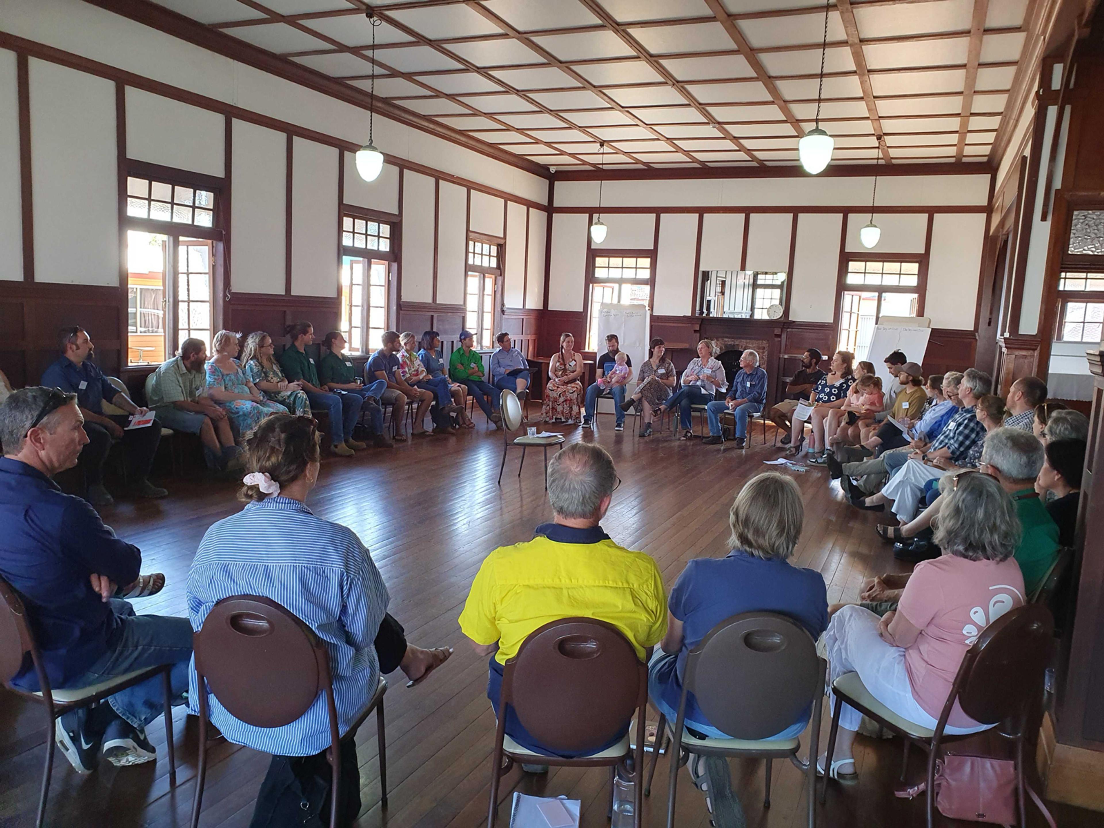 A group of approximately 50 people sitting in a circle in a hall.