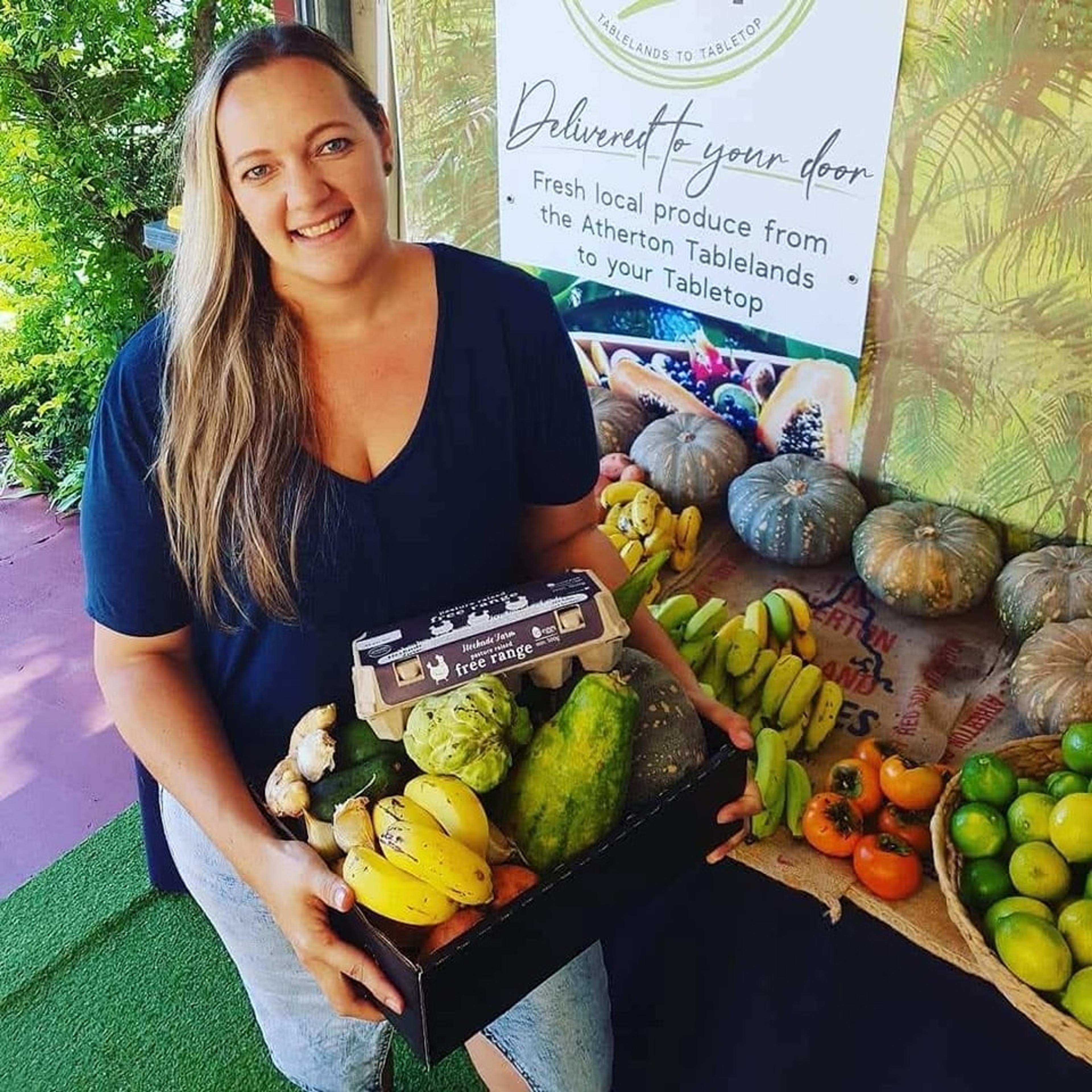 A smiling woman holding a box of fresh local produce including fruit, vegetables, eggs and more.