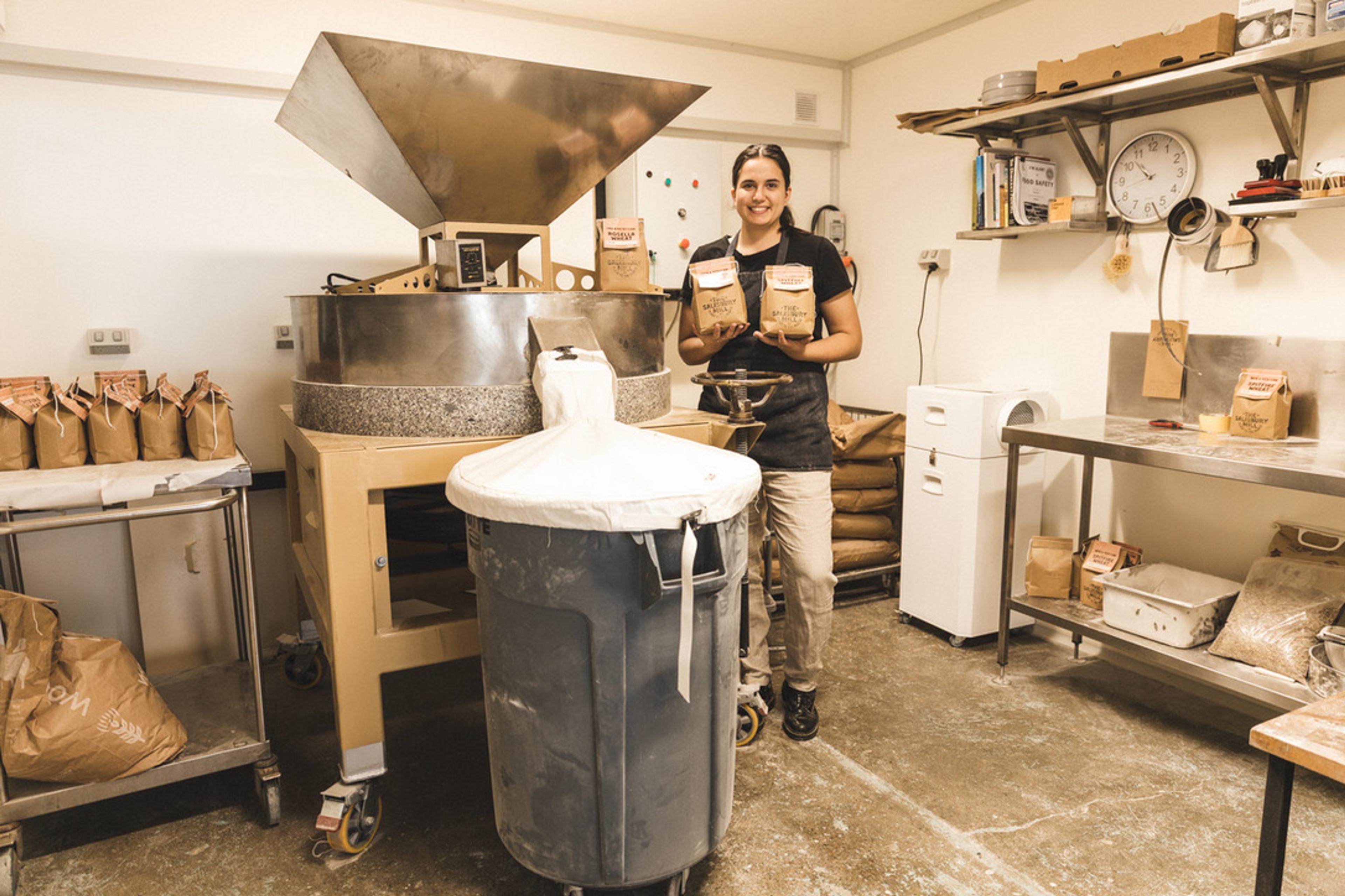 A person standing next to the mill with a bag of flour in each hand.