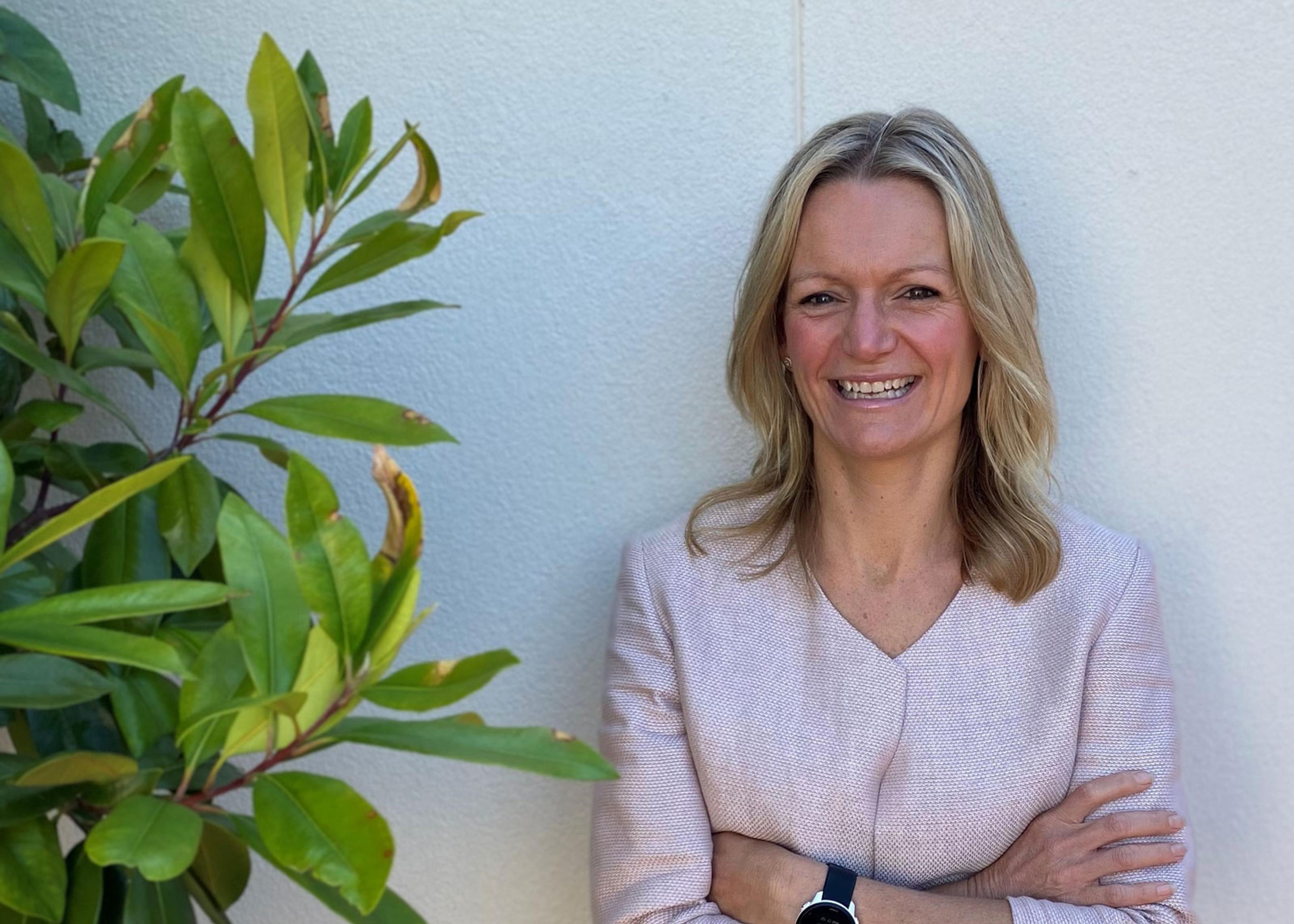 A photo of a smiling woman leaning against a wall. She is wearing a pink top. There is a plant to left of her.