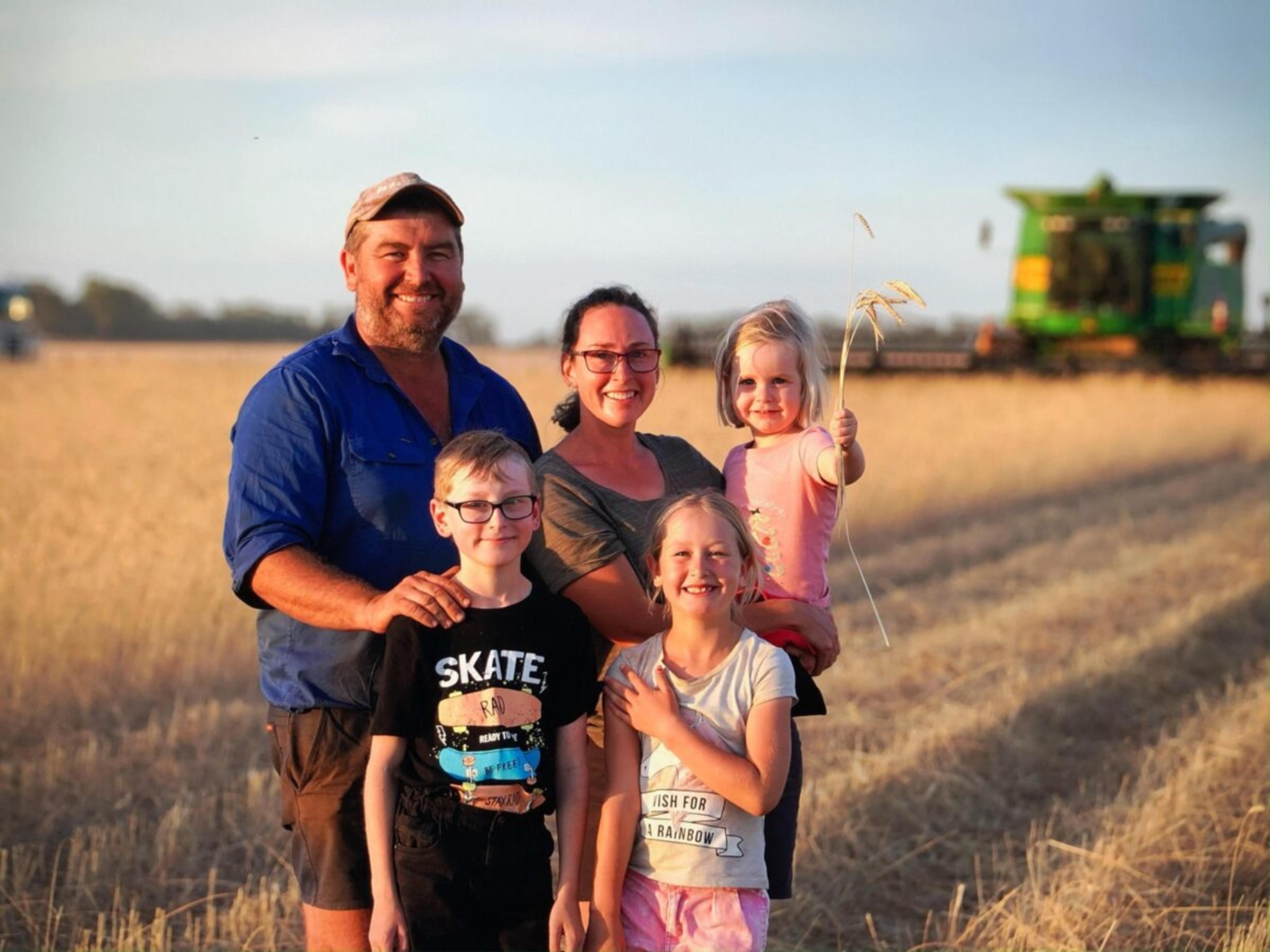 A farming family of 5, 2 adults and 3 children. They are standing in front of a paddock of wheat.