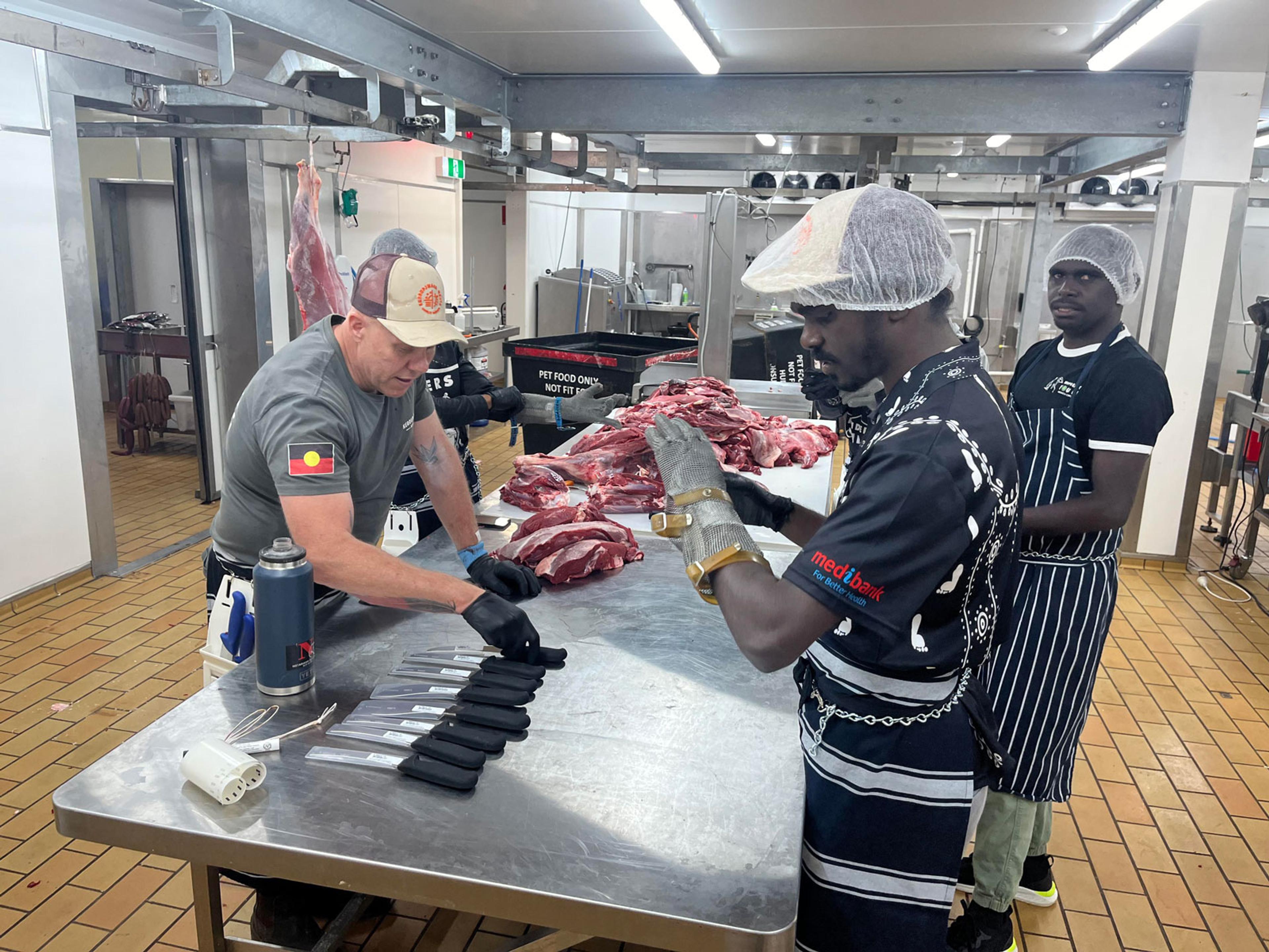3 men standing around a table looking at knives. There is a pile of red meat further along the table, ready to be butchered.