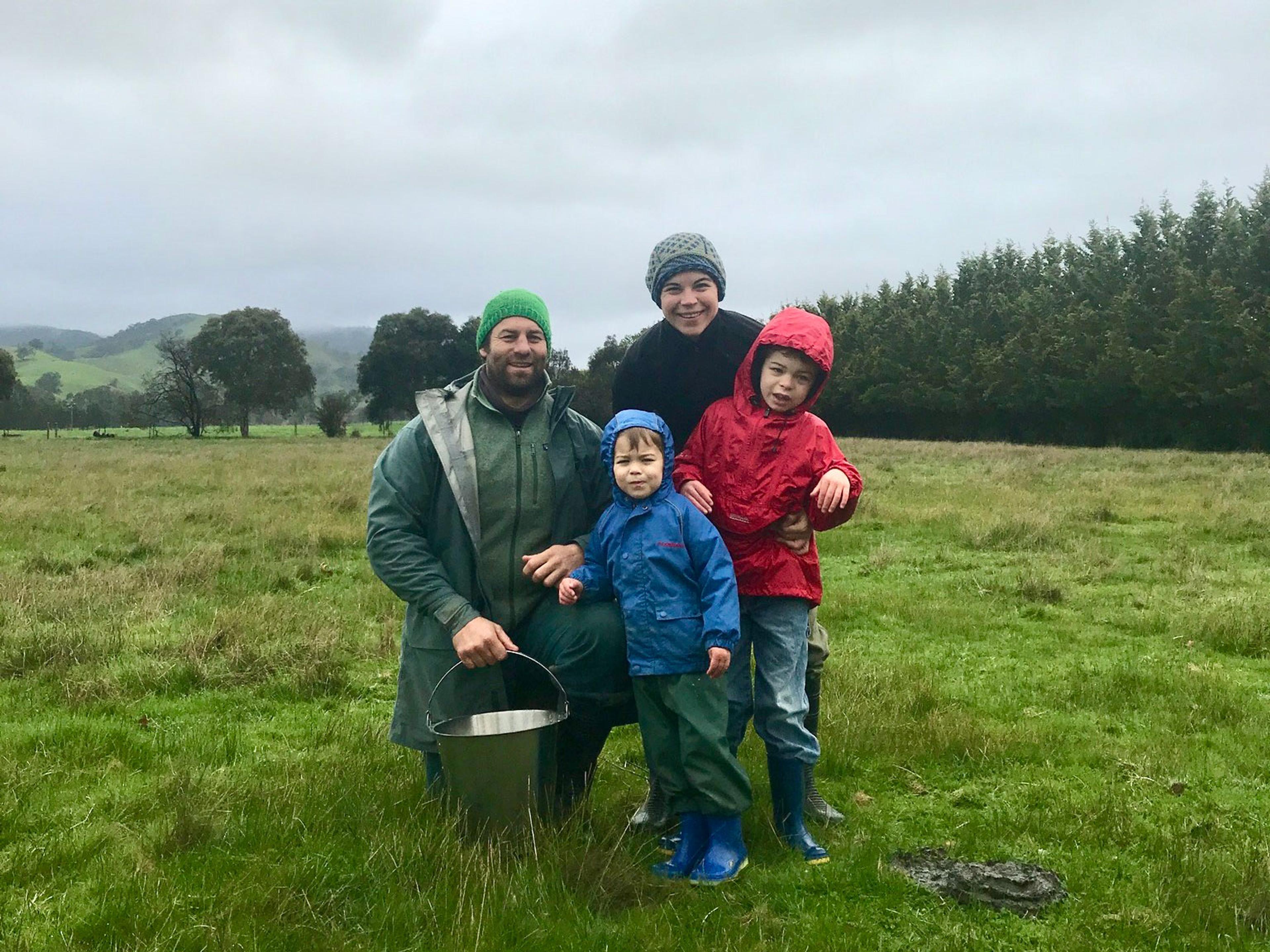 A couple and their two children posing for the camera. They're all rugged up with beanies and jackets. They're in the middle of a paddock with grey skies and a row of trees behind them. 