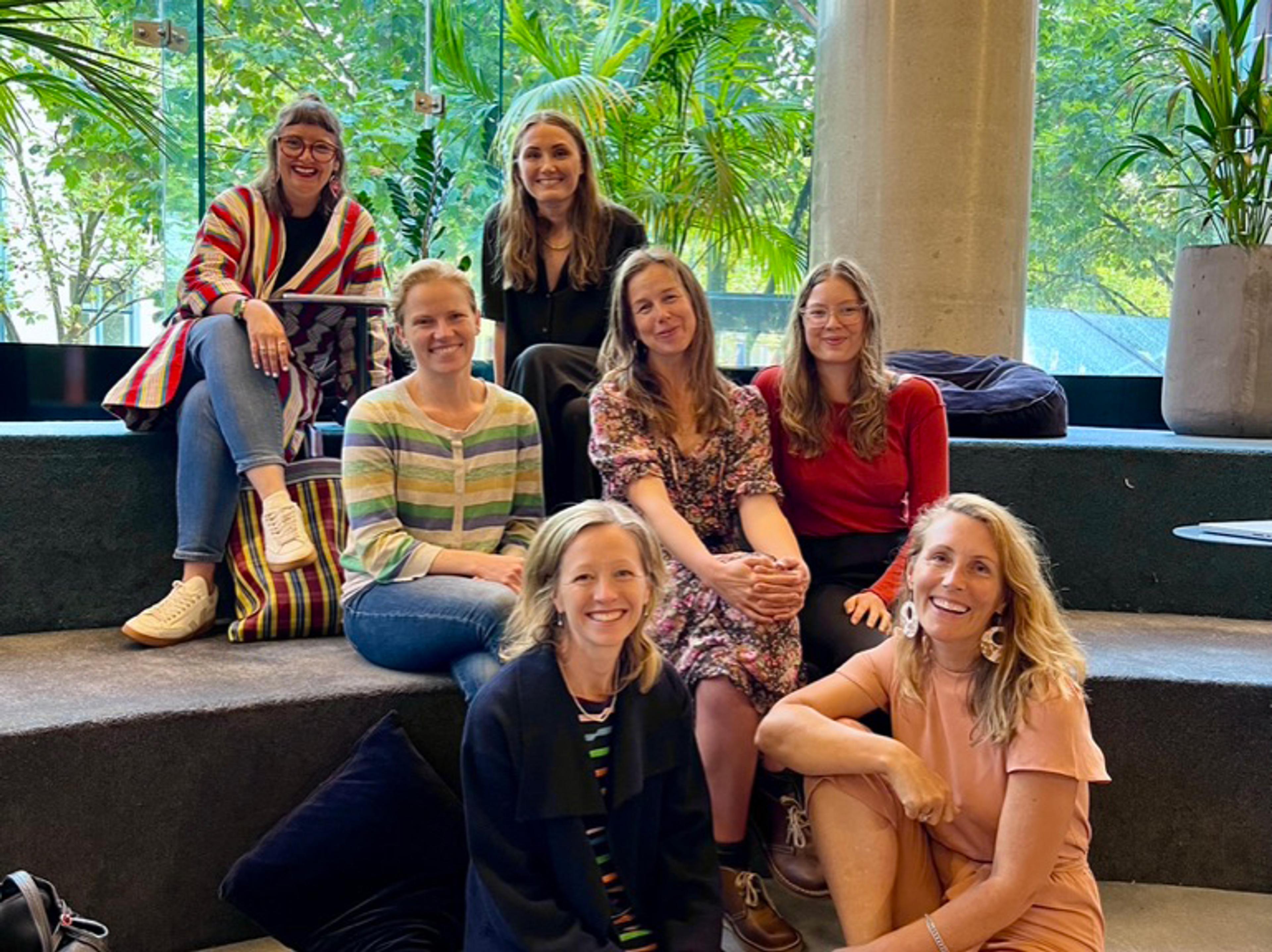Seven women sitting on stairs in a co-working space, all smiling at the camera.